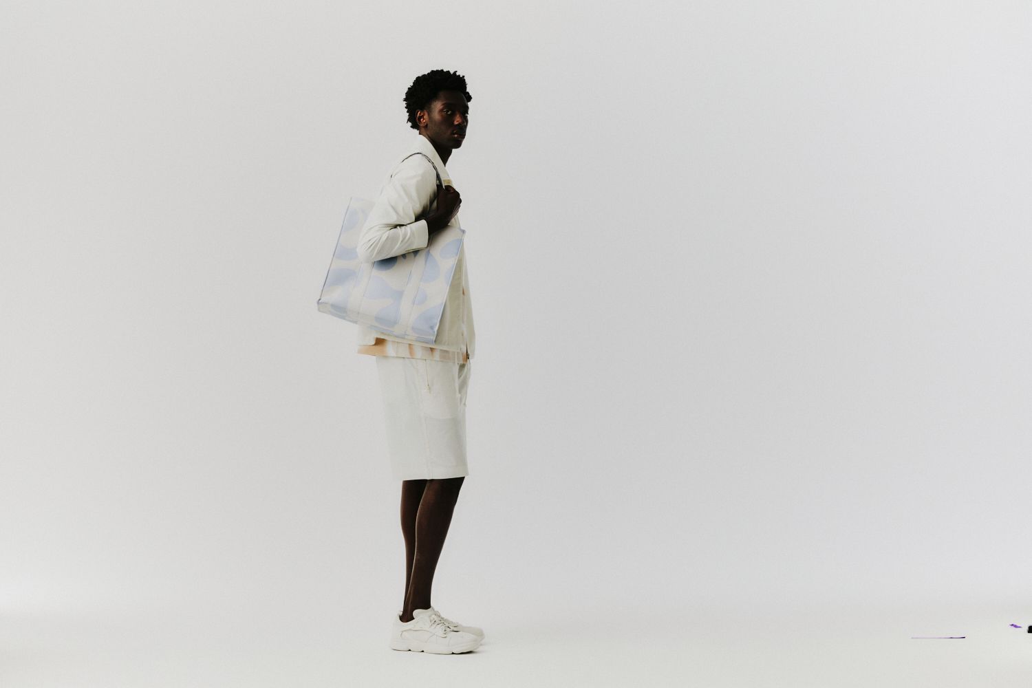 Person in cremefarbenem Outfit, die eine gemusterte Einkaufstasche trägt und vor einem weißen Hintergrund posiert.