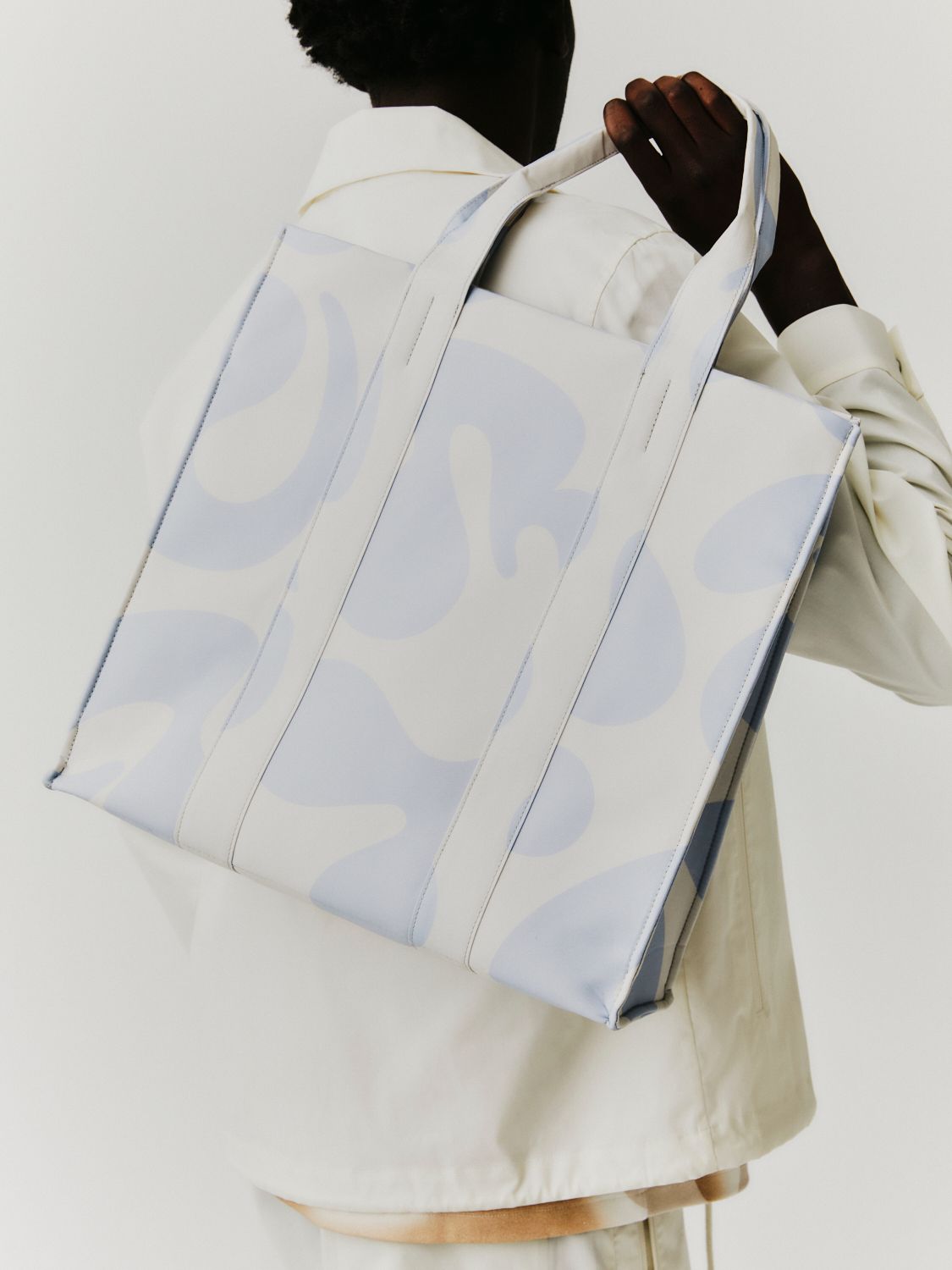 Person hält eine weiße Einkaufstasche mit hellblauen abstrakten Formen. Sie trägt ein weißes Hemd.