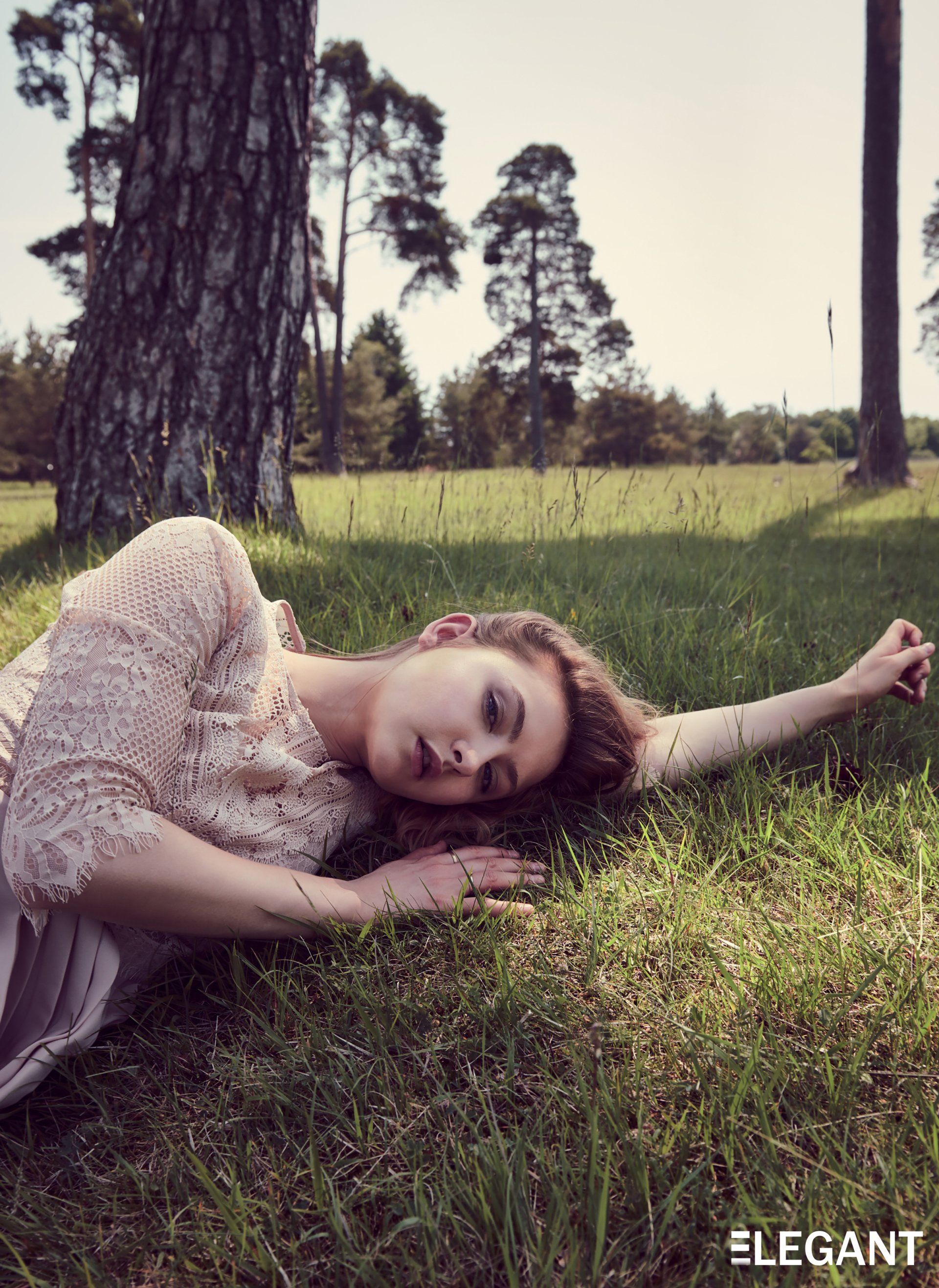 Frau im Kleid liegt im Gras unter einem Baum und blickt im Sonnenlicht in die Kamera.