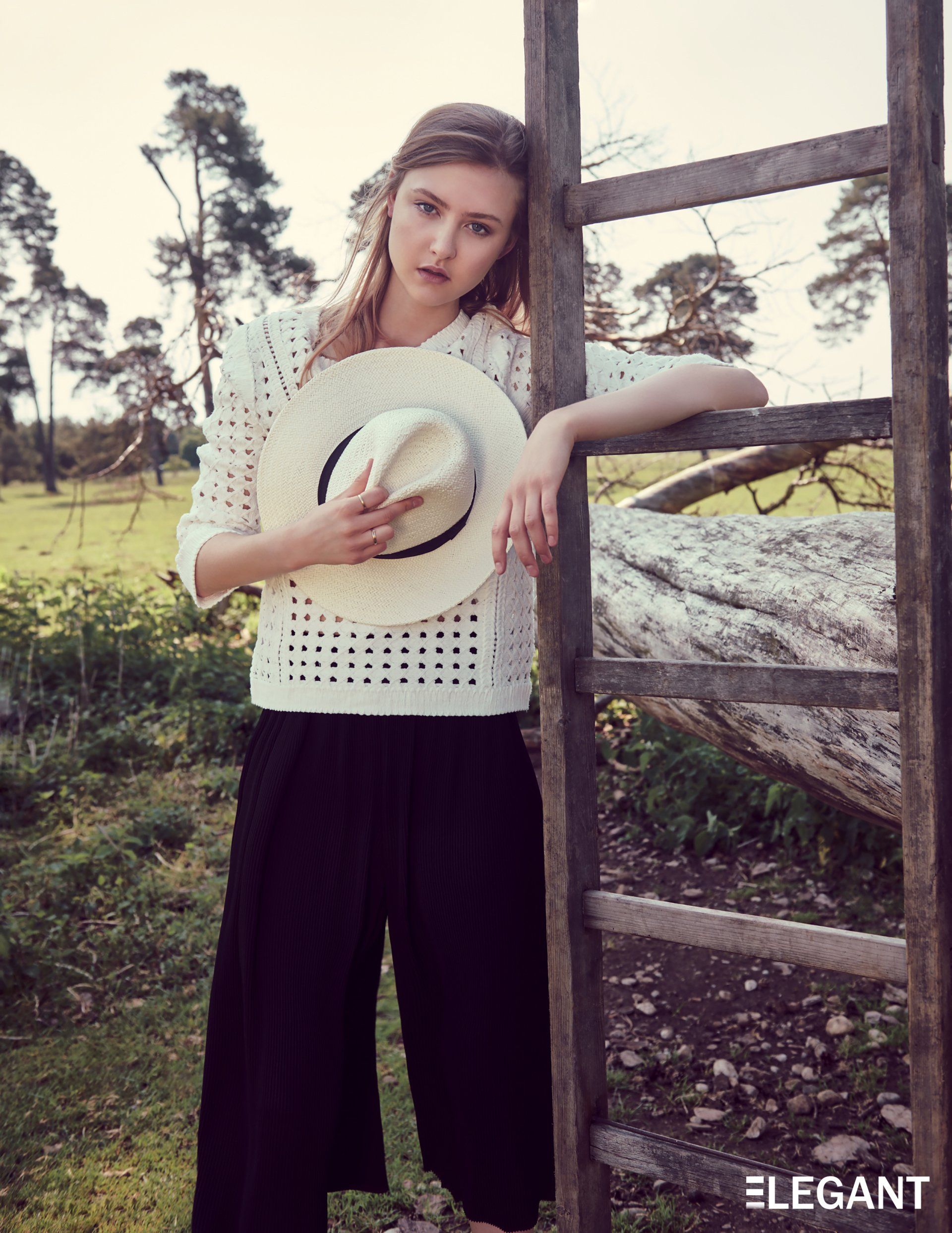 Frau mit weißem Oberteil und schwarzer Hose, Hut in der Hand, lehnt im Freien an einem Holzzaun.