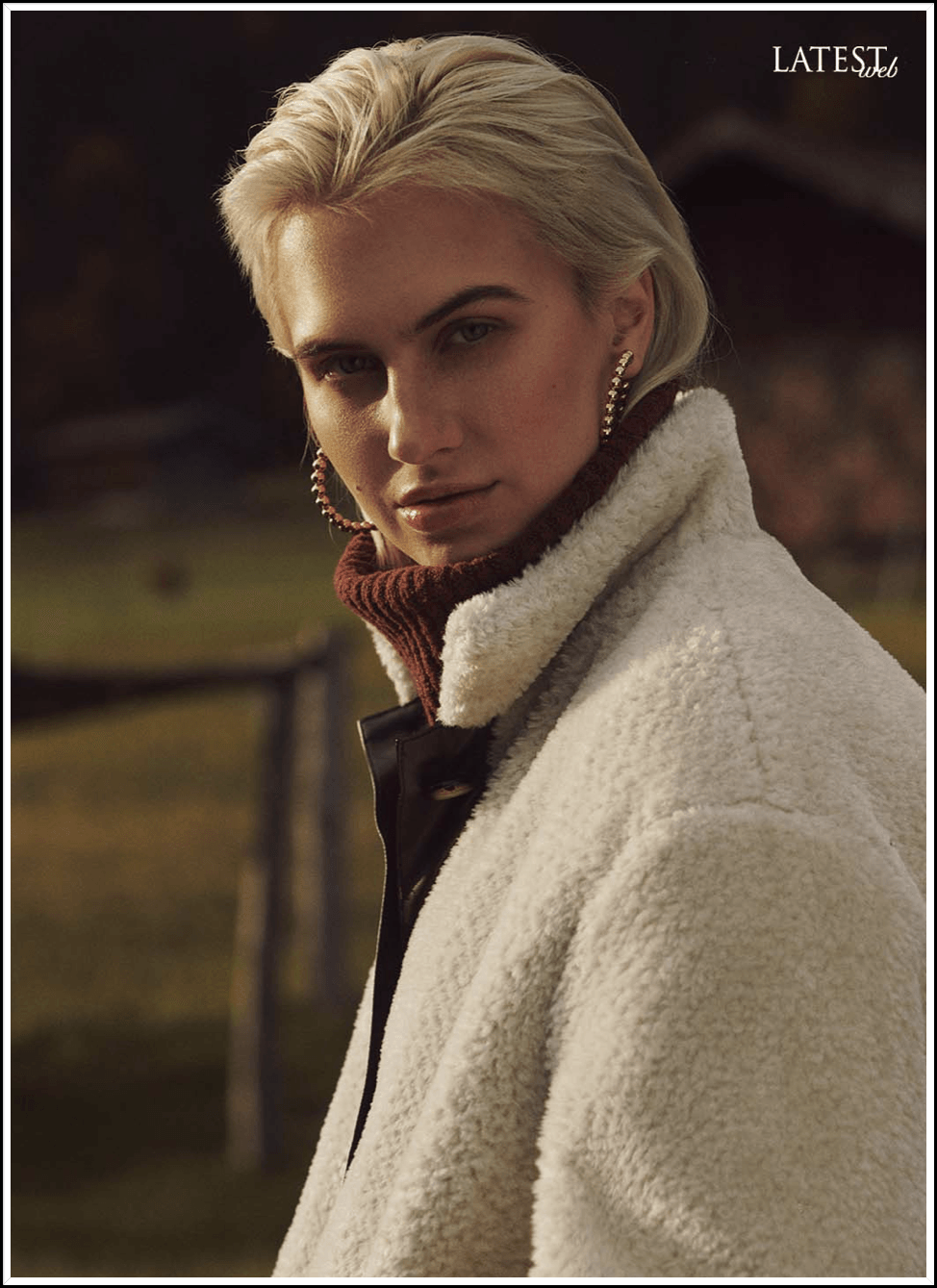 Frau mit blondem Haar, weißem Mantel und Creolen, blickt den Betrachter an.