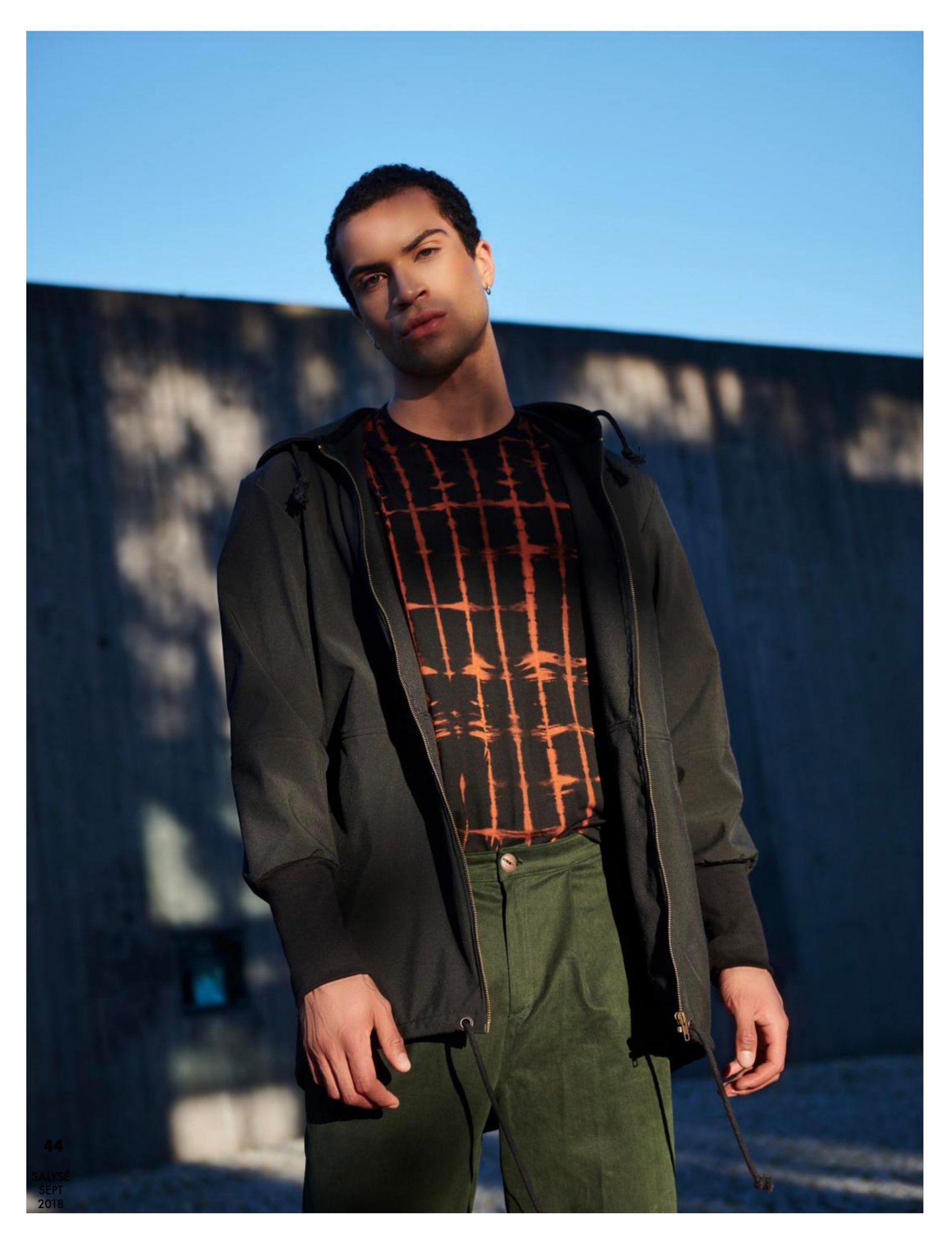 Mann in olivgrüner Hose, schwarzer Jacke über gemustertem Hemd, posiert im Freien vor blauem Himmel.