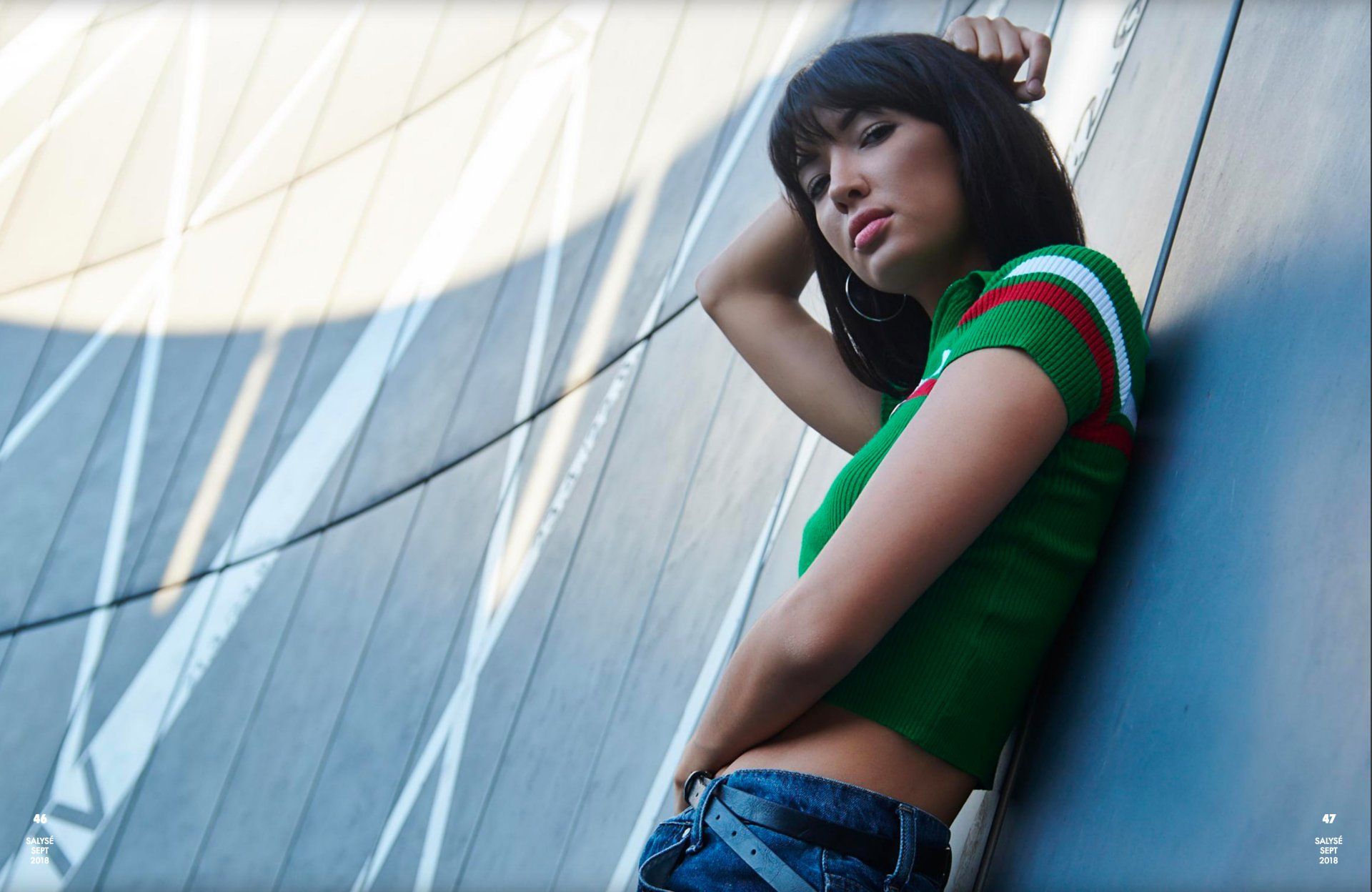 Frau lehnt an einer strukturierten Wand, trägt grünes Oberteil und Jeans und blickt in die Kamera.