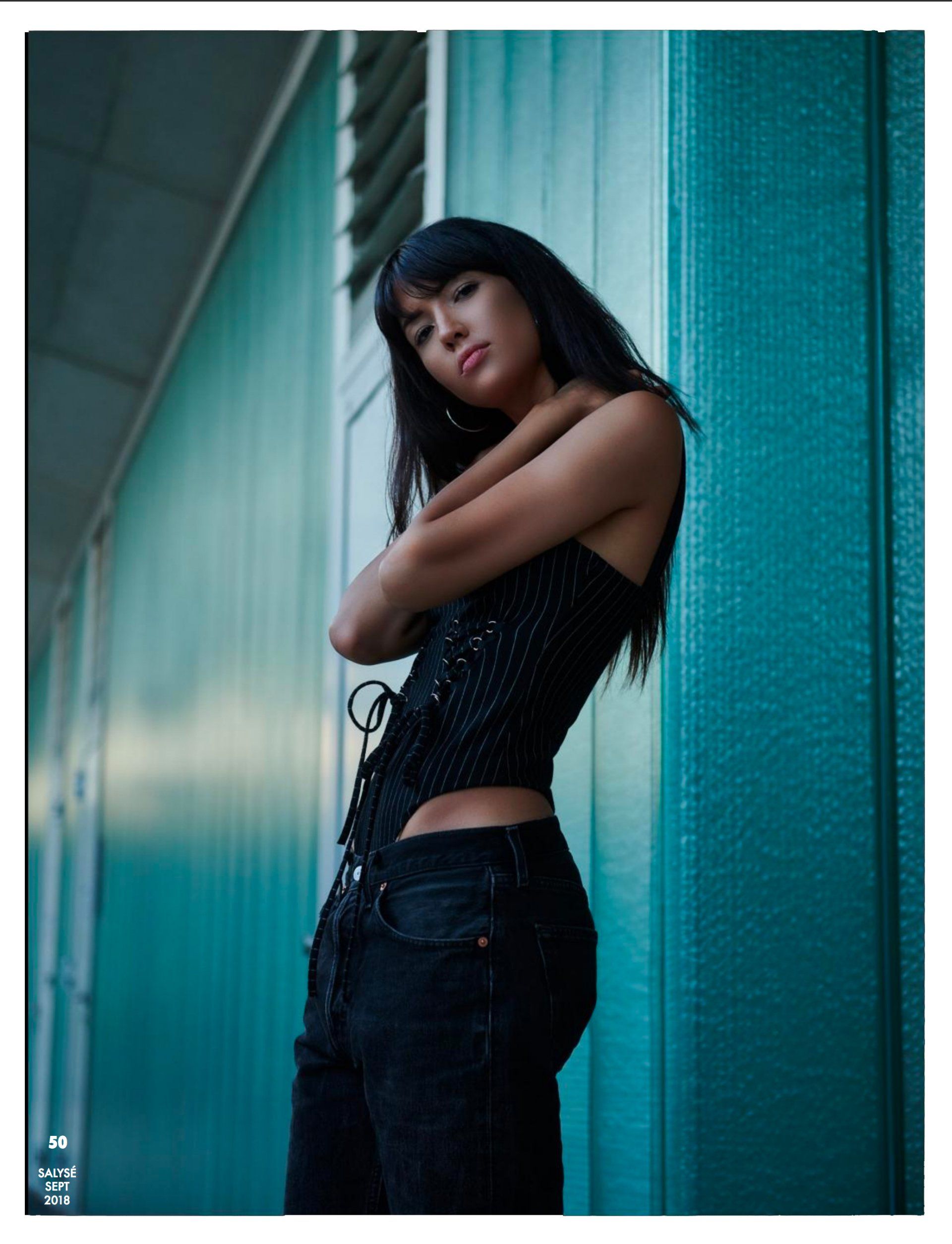 Frau in schwarzem Korsett und Jeans, Arme verschränkt, posiert vor einer blaugrünen Wand.