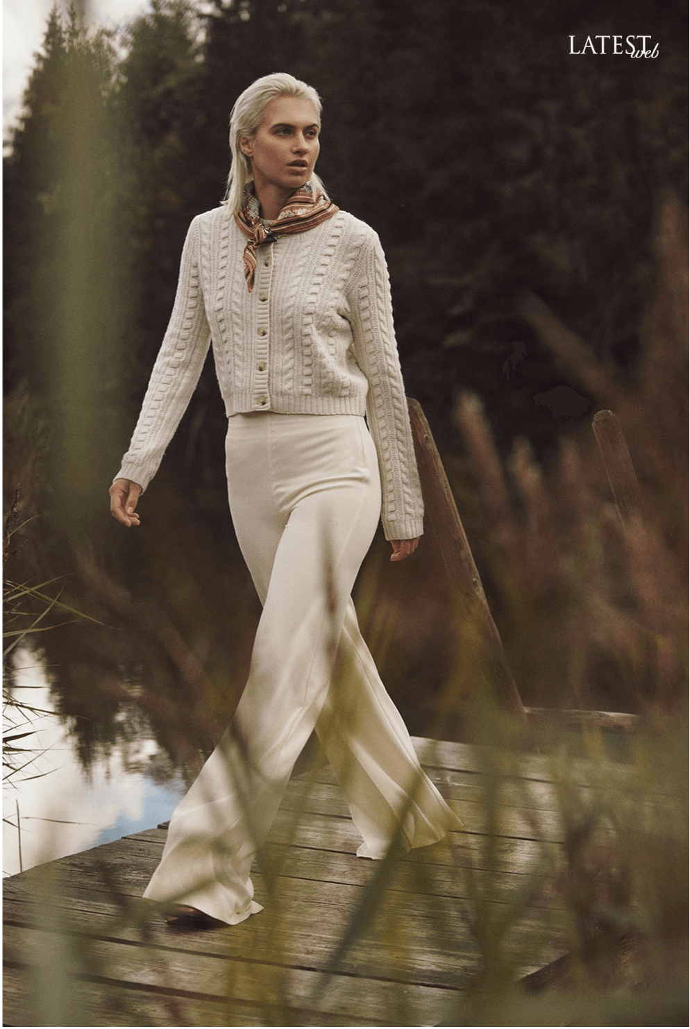 Frau in weißer Strickjacke und Hose geht auf einem Holzsteg; Umgebung im Freien.