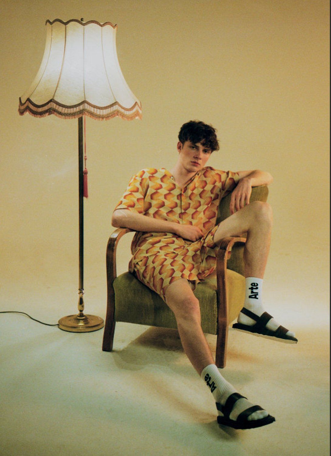 Mann im Outfit mit Ananas-Print sitzt im Sessel, Lampe in der Nähe. Beiger Hintergrund.