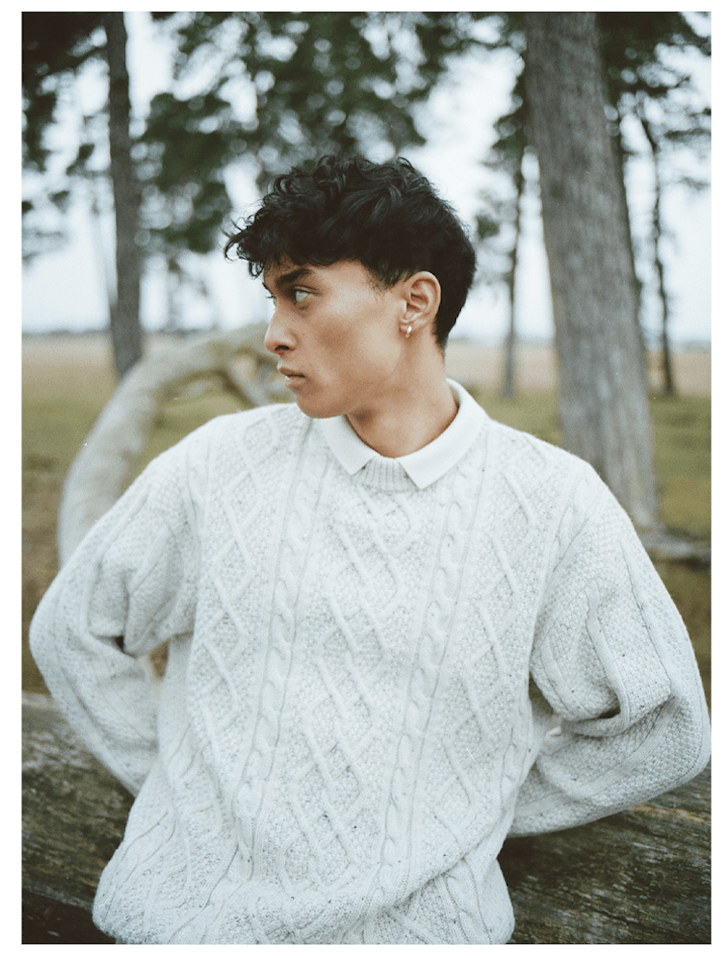 Mann mit lockigem Haar und cremefarbenem Pullover, der im Wald auf einem Baumstamm lehnt und wegschaut.