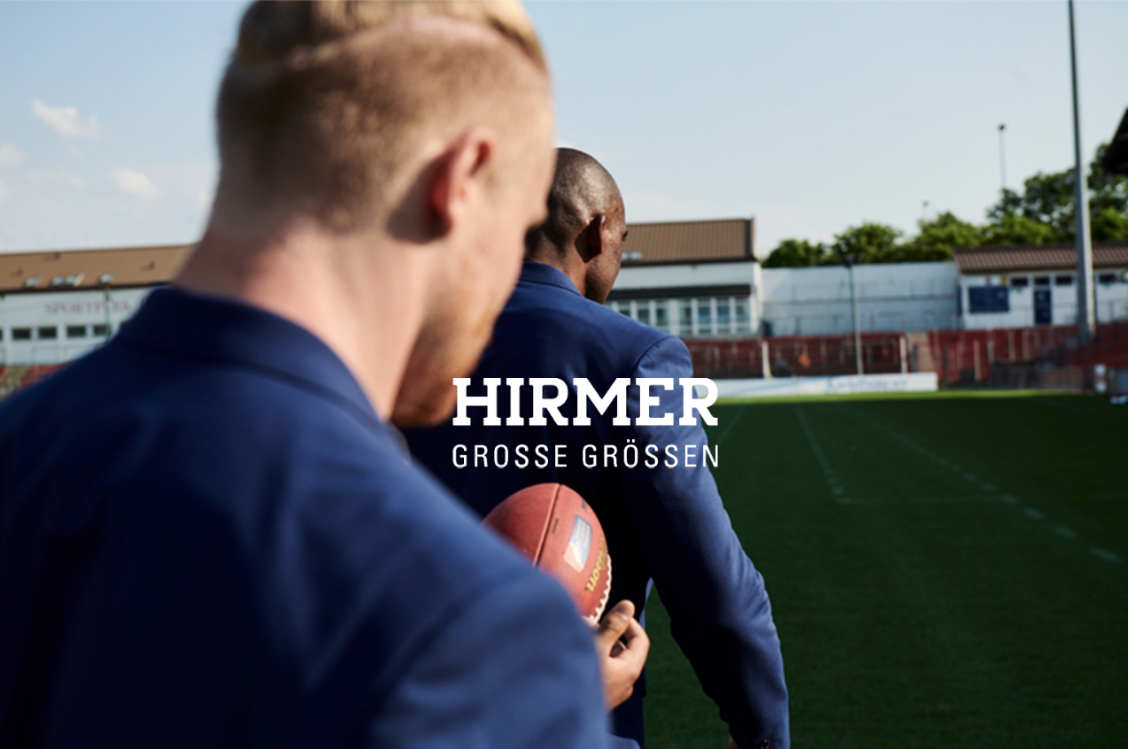 Zwei Männer in Anzügen auf einem Fußballfeld, einer hält einen Ball. HIRMER-Logo.