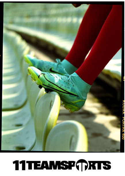 Füße eines Fußballspielers mit blaugrünen Stollen und roten Socken ruhen auf Stadionsitzen; „11 TEAMSPORTS“-Logo.