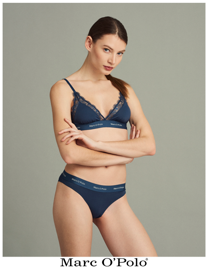 Frau in blauer Unterwäsche, die Arme verschränkt, blickt den Betrachter vor grauem Hintergrund an.