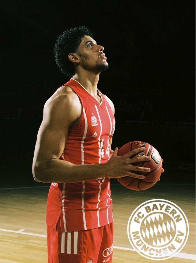 Basketballspieler hält einen Ball, trägt eine rote Uniform des FC Bayern München und blickt nach oben.