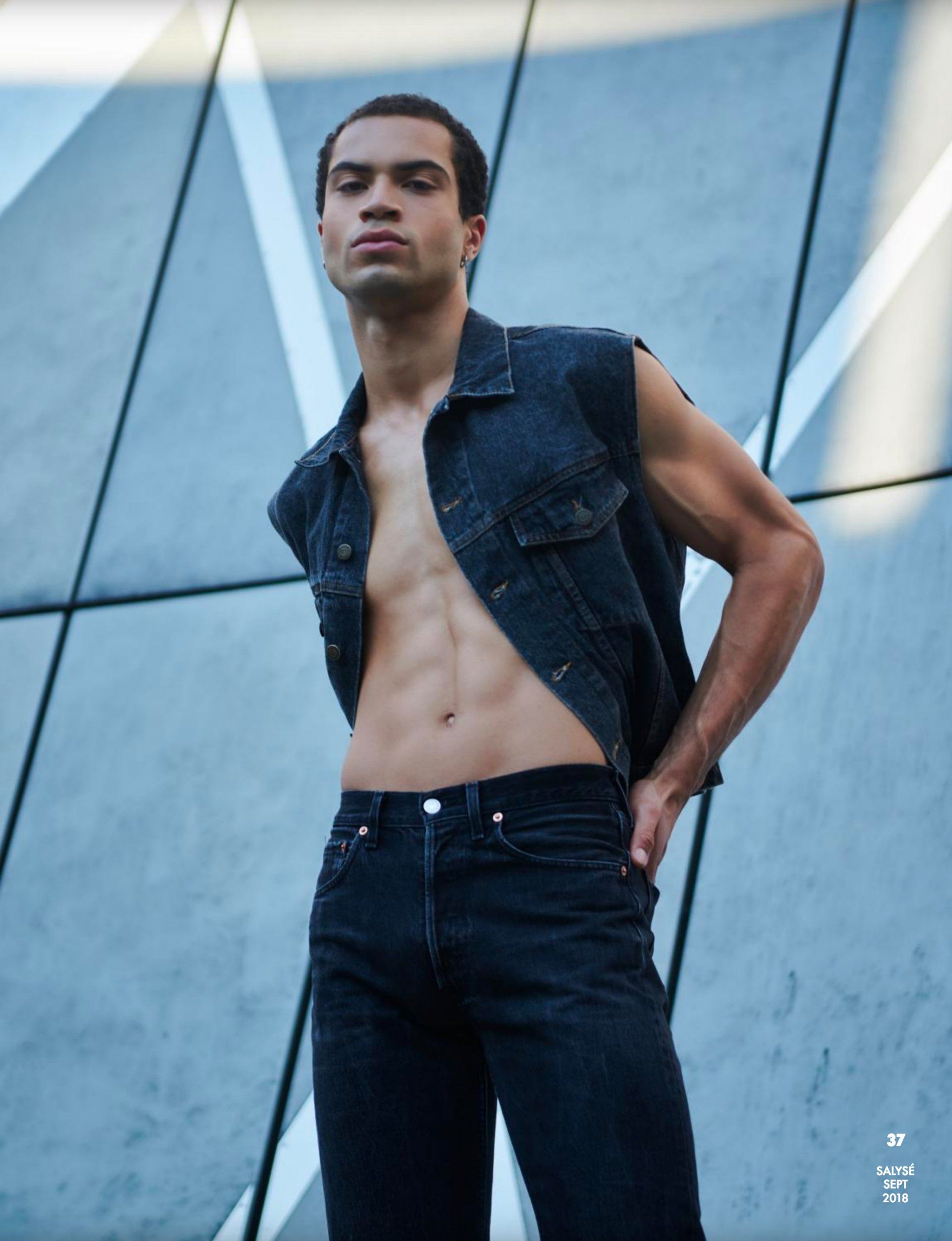 Mann in Jeansweste und Jeans steht mit der Hand in der Tasche vor einem modernen Gebäude.