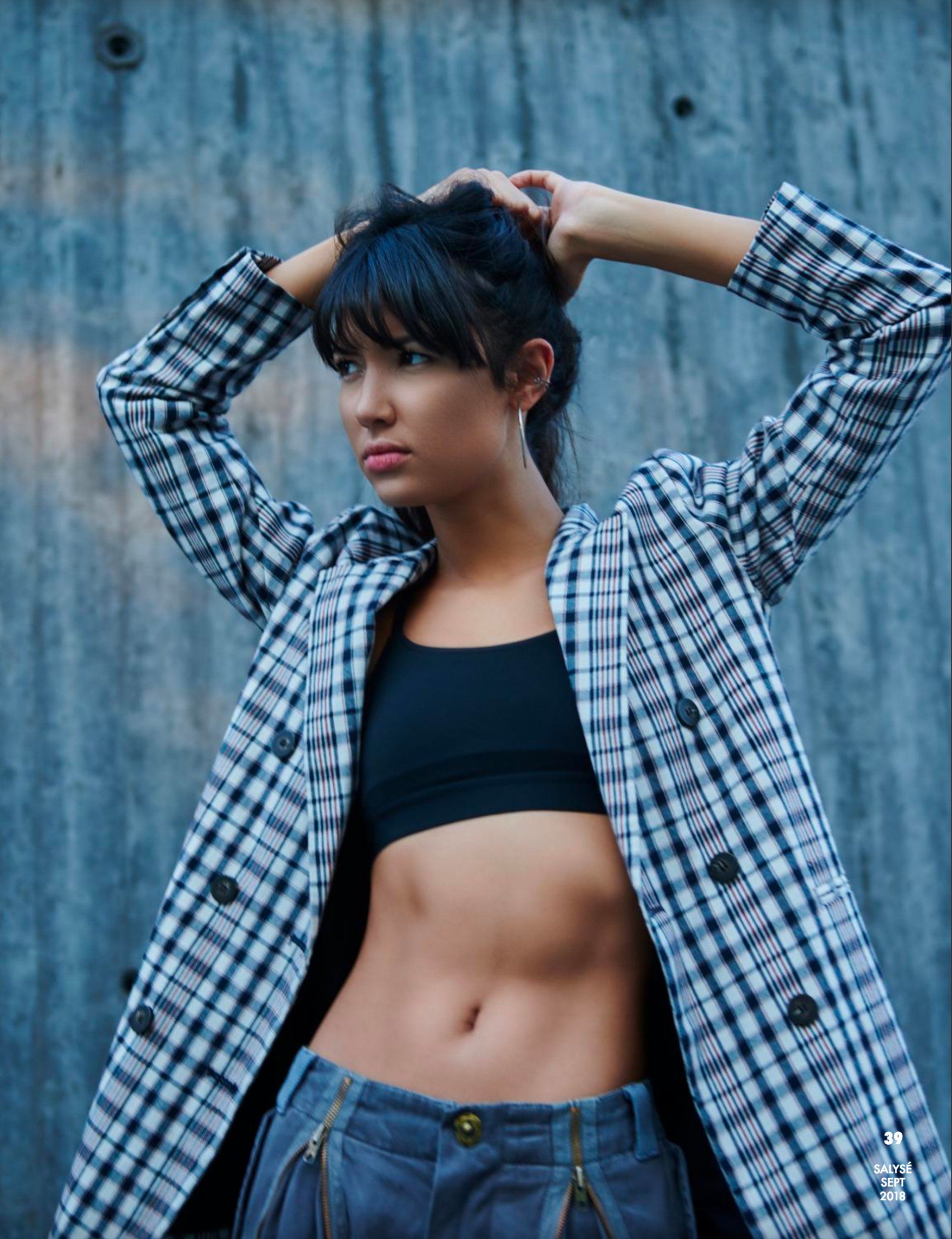 Frau in kariertem Blazer, schwarzem bauchfreiem Top und grauer Hose posiert neben einer blauen Wand, die Hände im Haar.