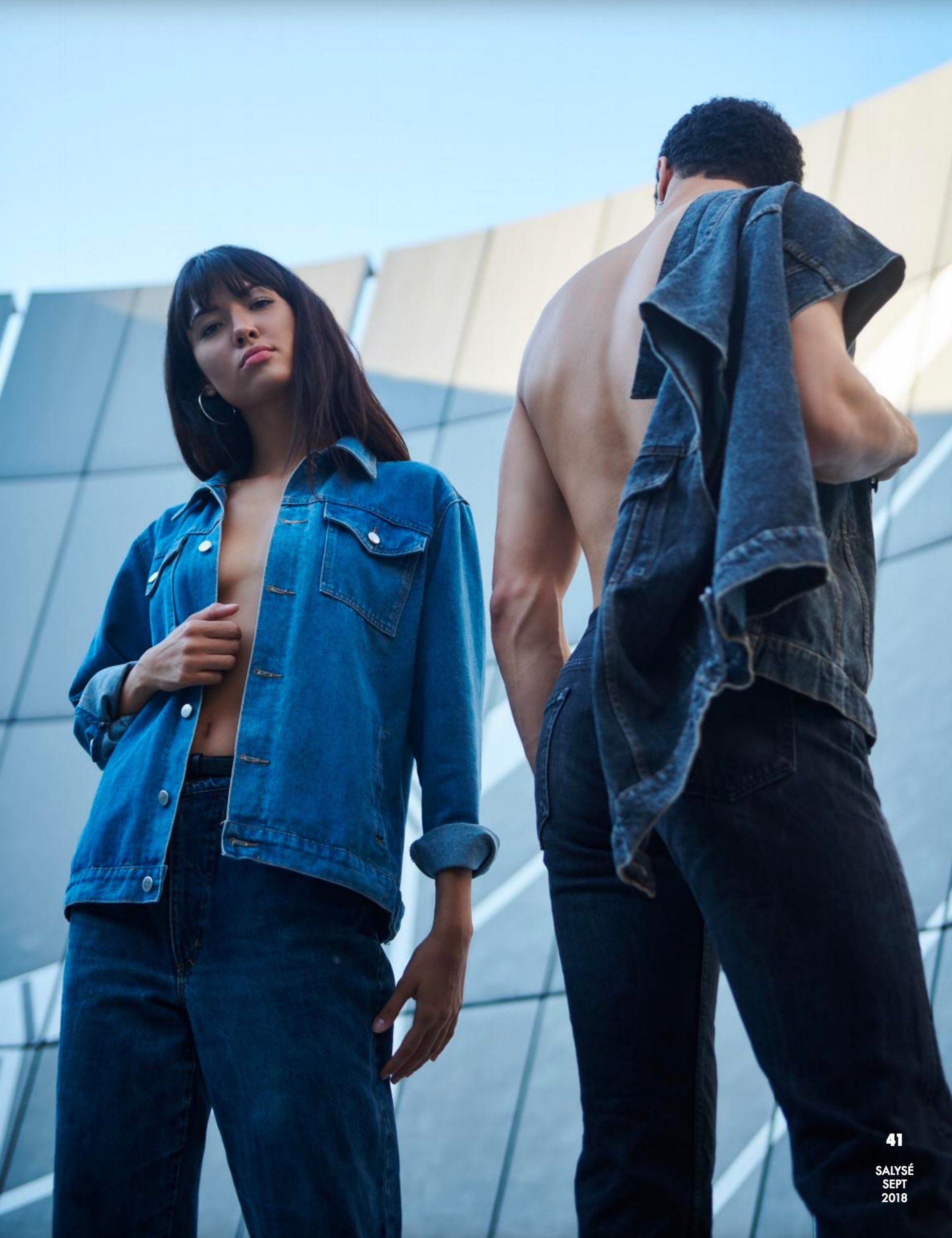 Frau in blauer Jeansjacke und -hose und Mann mit Jeansjacke stehen draußen.