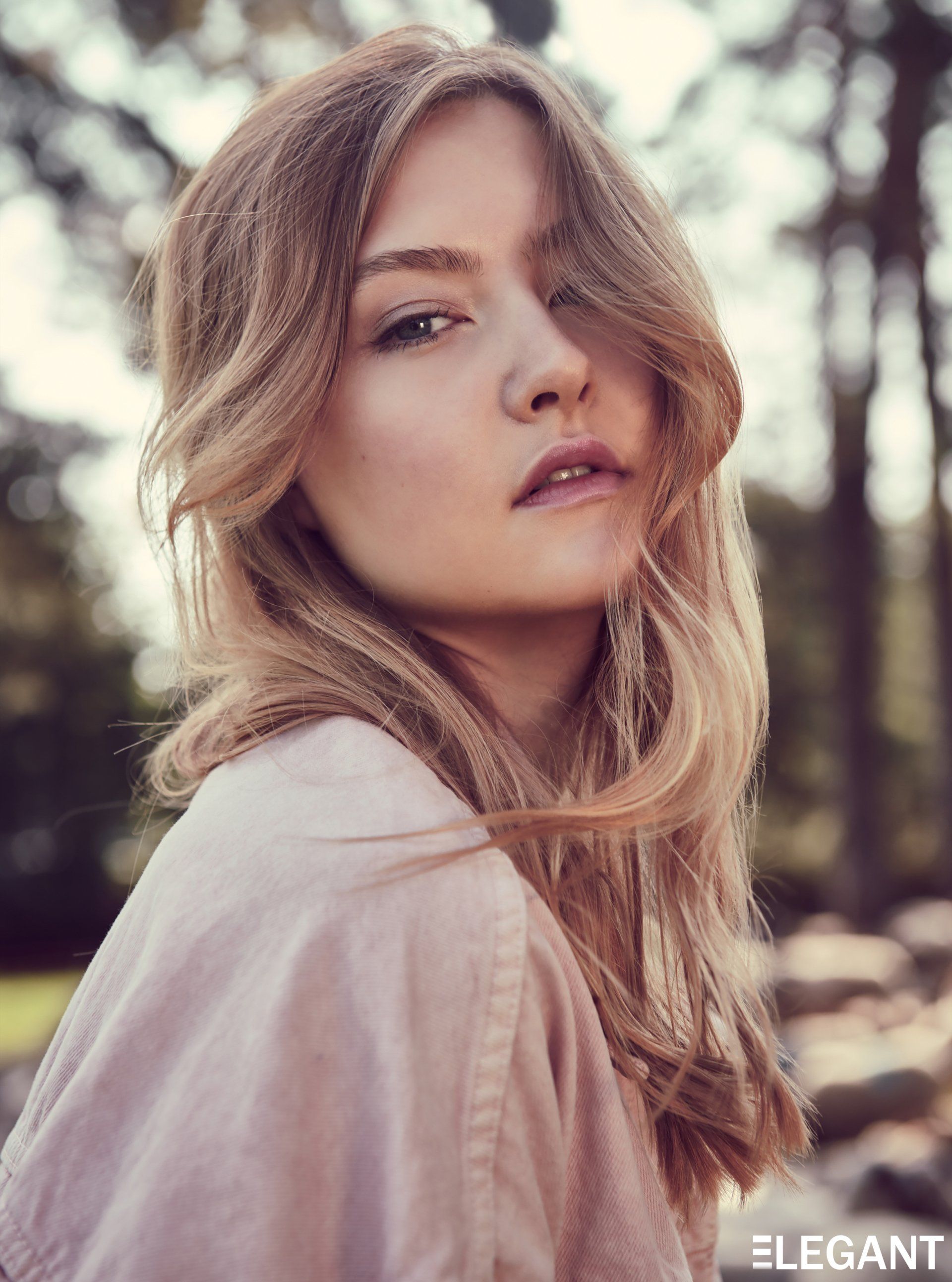 Blonde Frau mit sanftem Gesichtsausdruck, trägt ein rosa Hemd, im Freien.