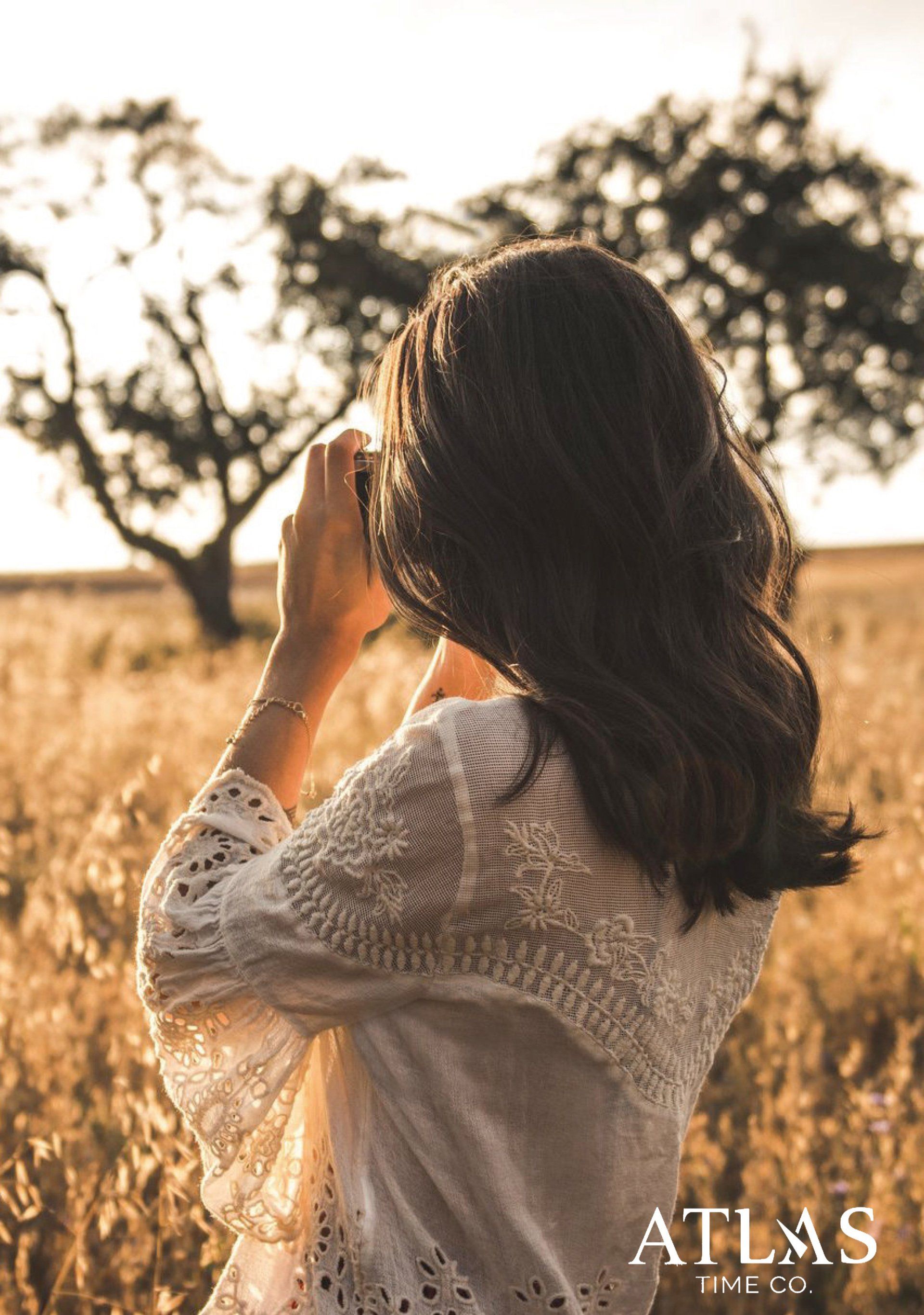 Frau macht ein Foto in einem Feld aus goldenem Weizen, im Hintergrund ein Baum.