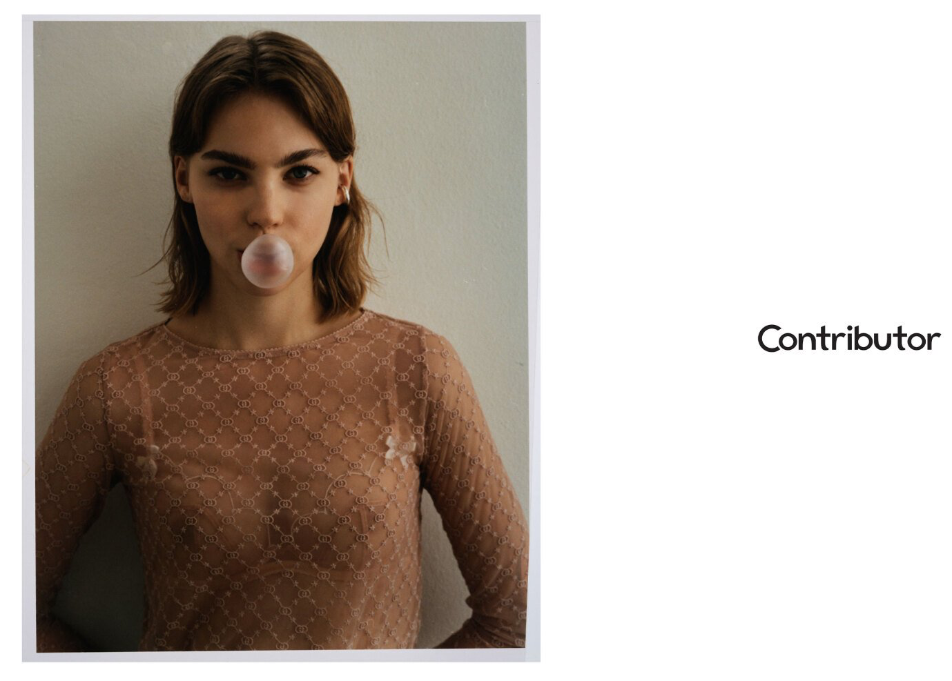 Frau in einem braunen Netzoberteil bläst vor einer weißen Wand eine Kaugummiblase.