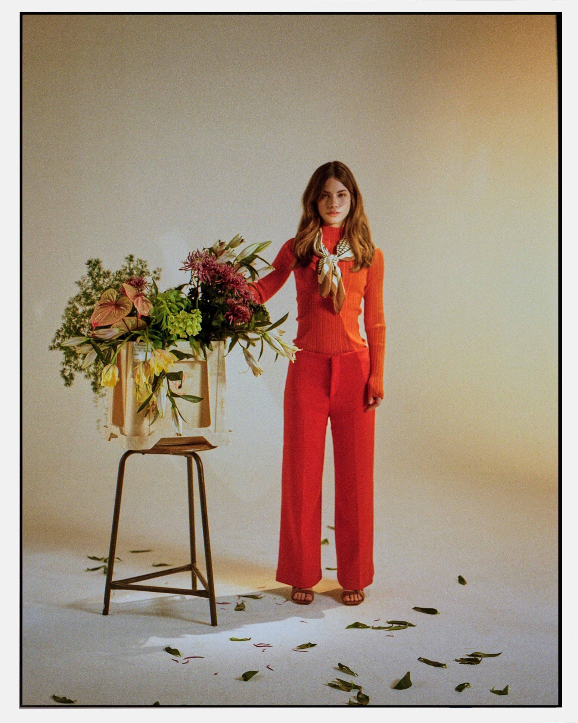 Frau im roten Overall steht neben einem Hocker mit einem großen Blumenarrangement. Blätter auf dem Boden.