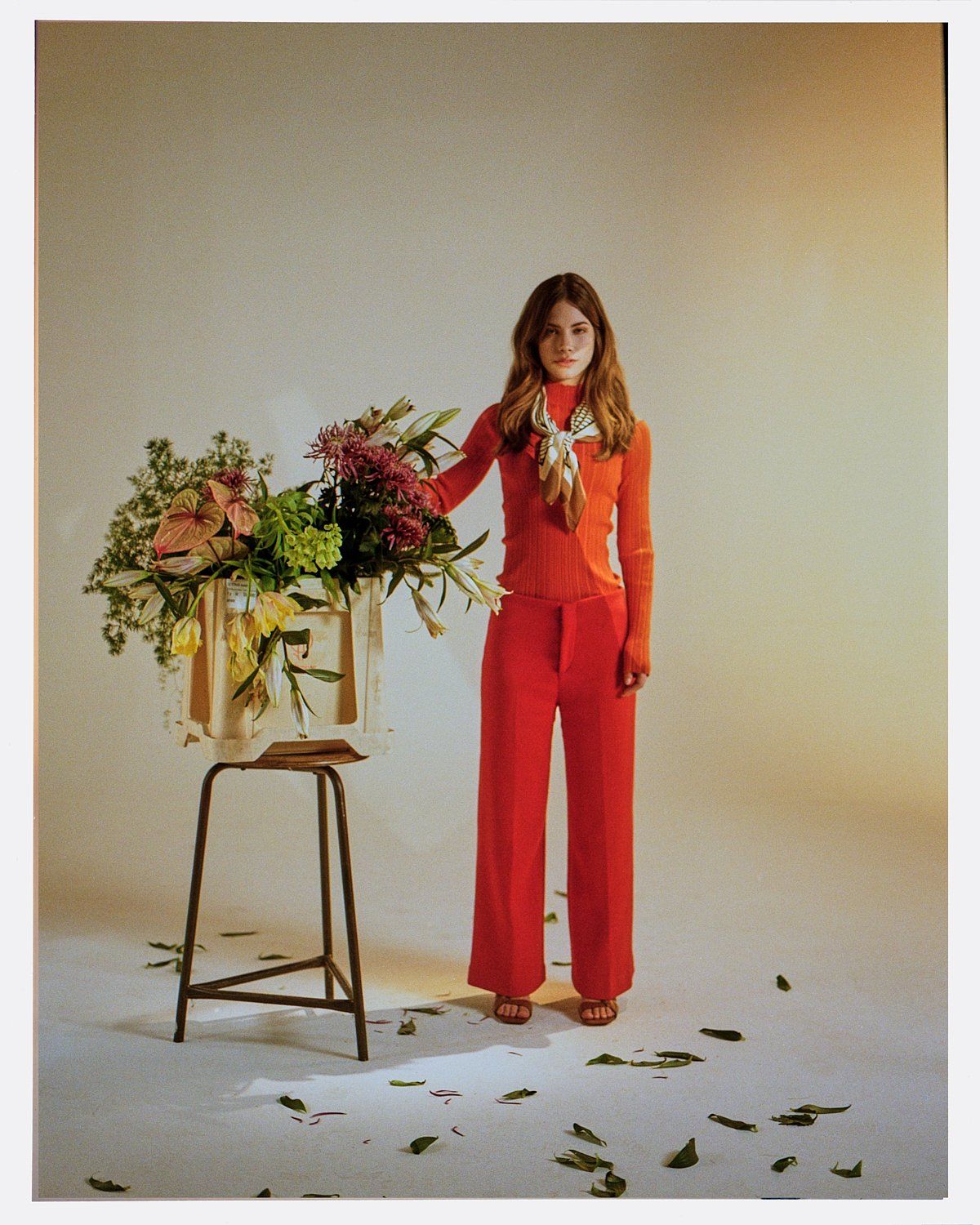 Frau im roten Hosenanzug steht neben Hocker mit Blumenarrangement, auf dem Boden sind Blätter verstreut.