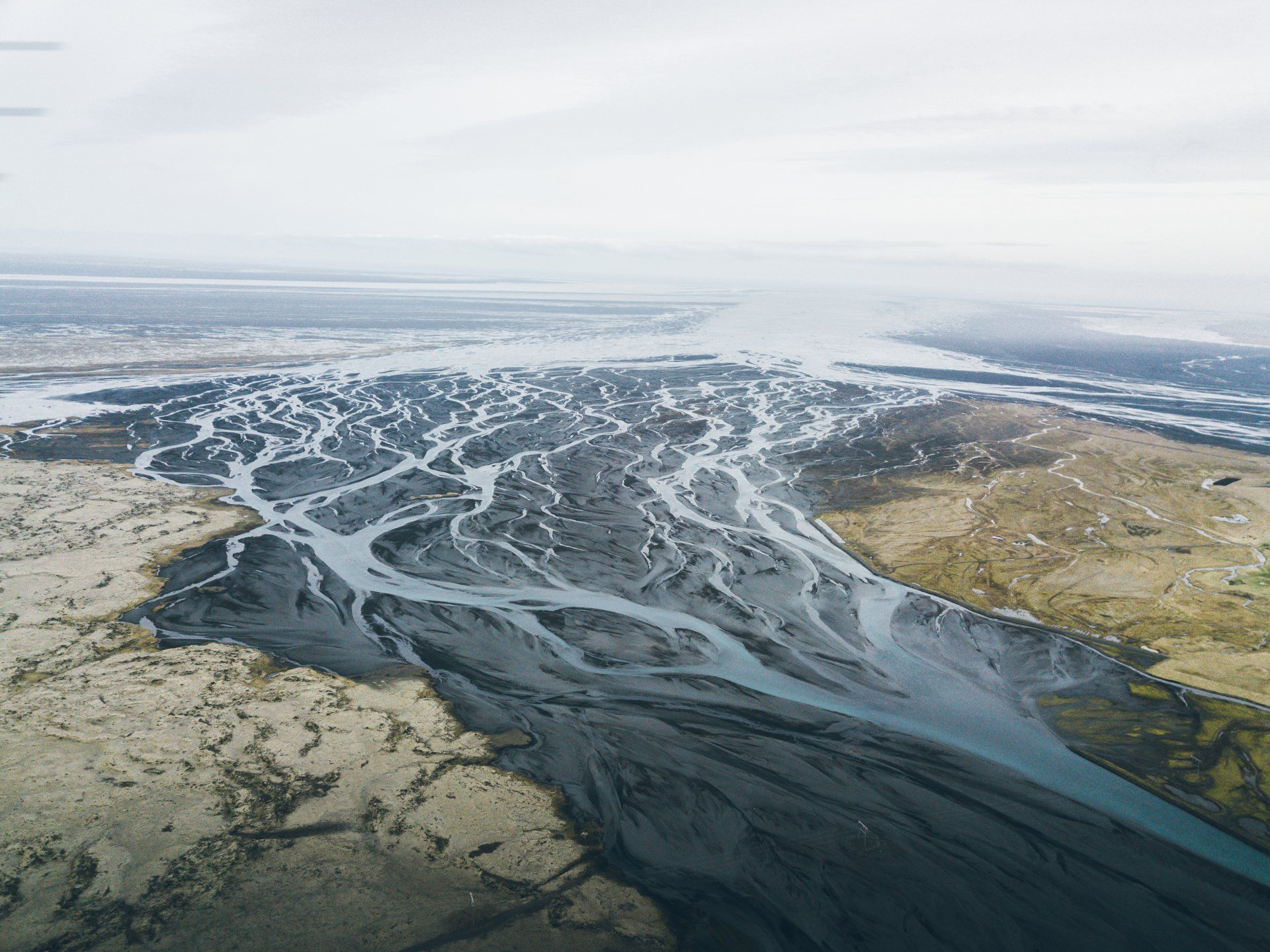 Verzweigter Gletscherfluss, der unter einem bewölkten Himmel über eine flache, sandige Ebene fließt.