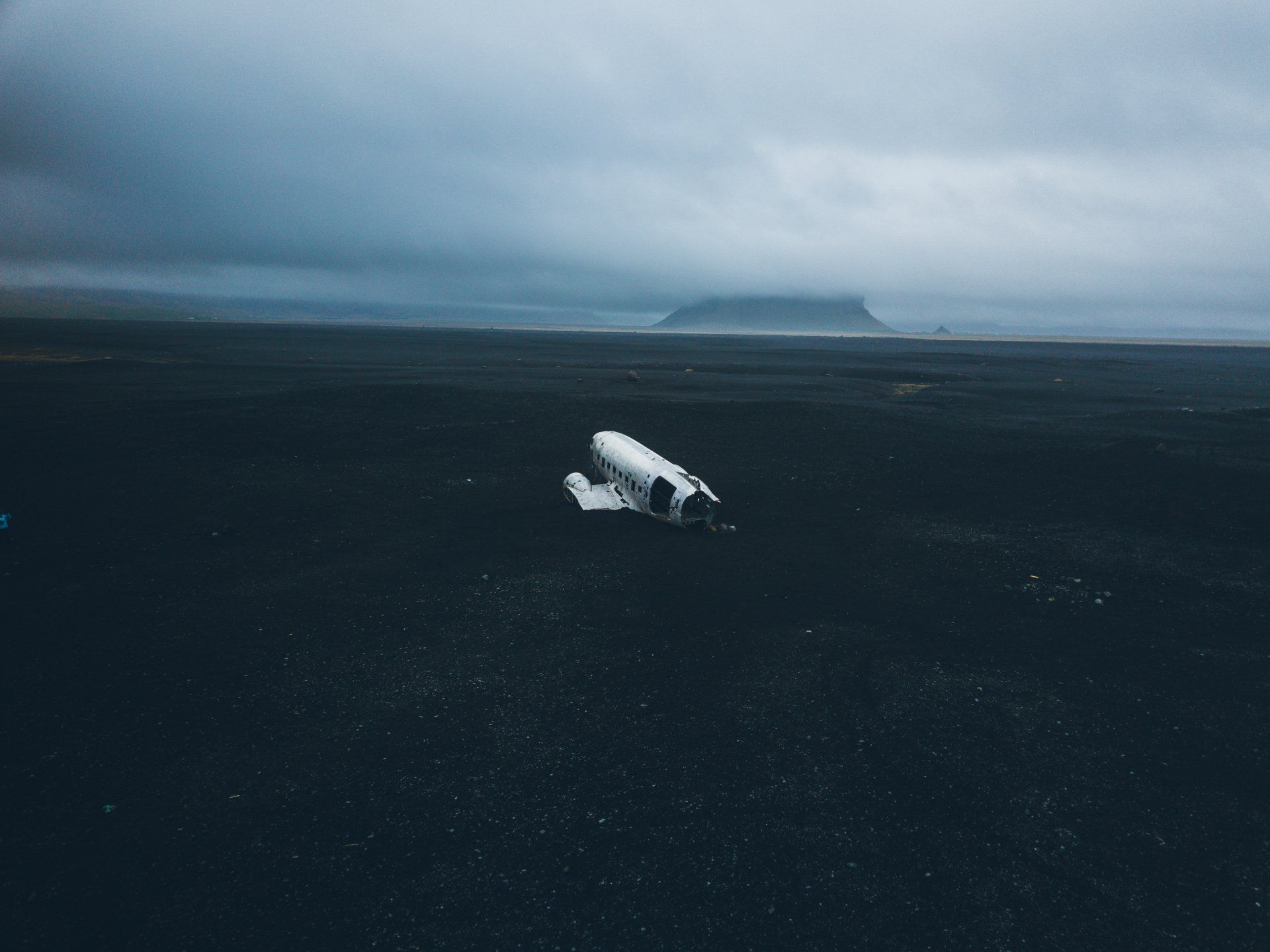 Wrack eines weißen Flugzeugs an einem schwarzen Sandstrand unter einem bewölkten Himmel, mit einem Berg in der Ferne.