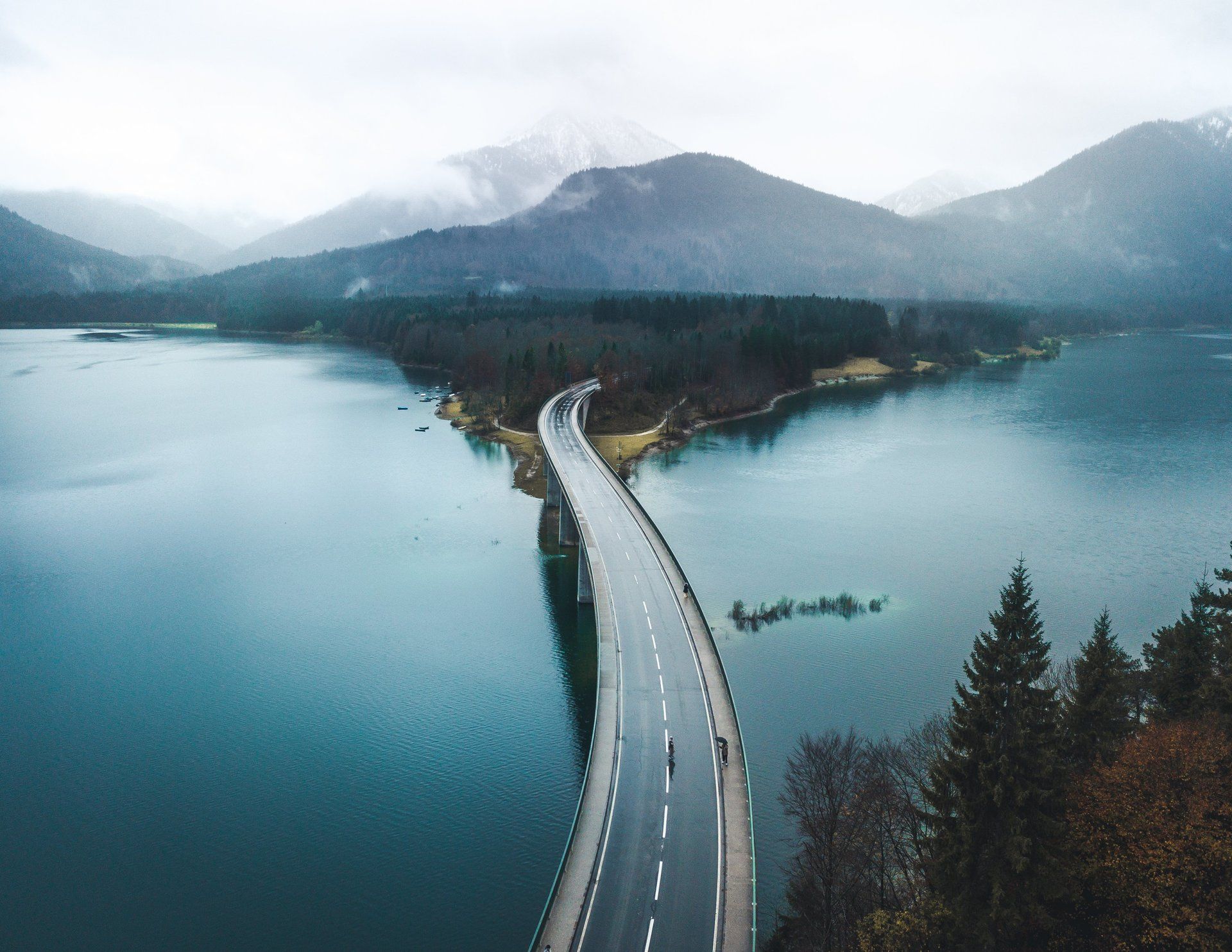 Brücke über einen blauen See, umgeben von Bergen und immergrünen Bäumen an einem bewölkten Tag.