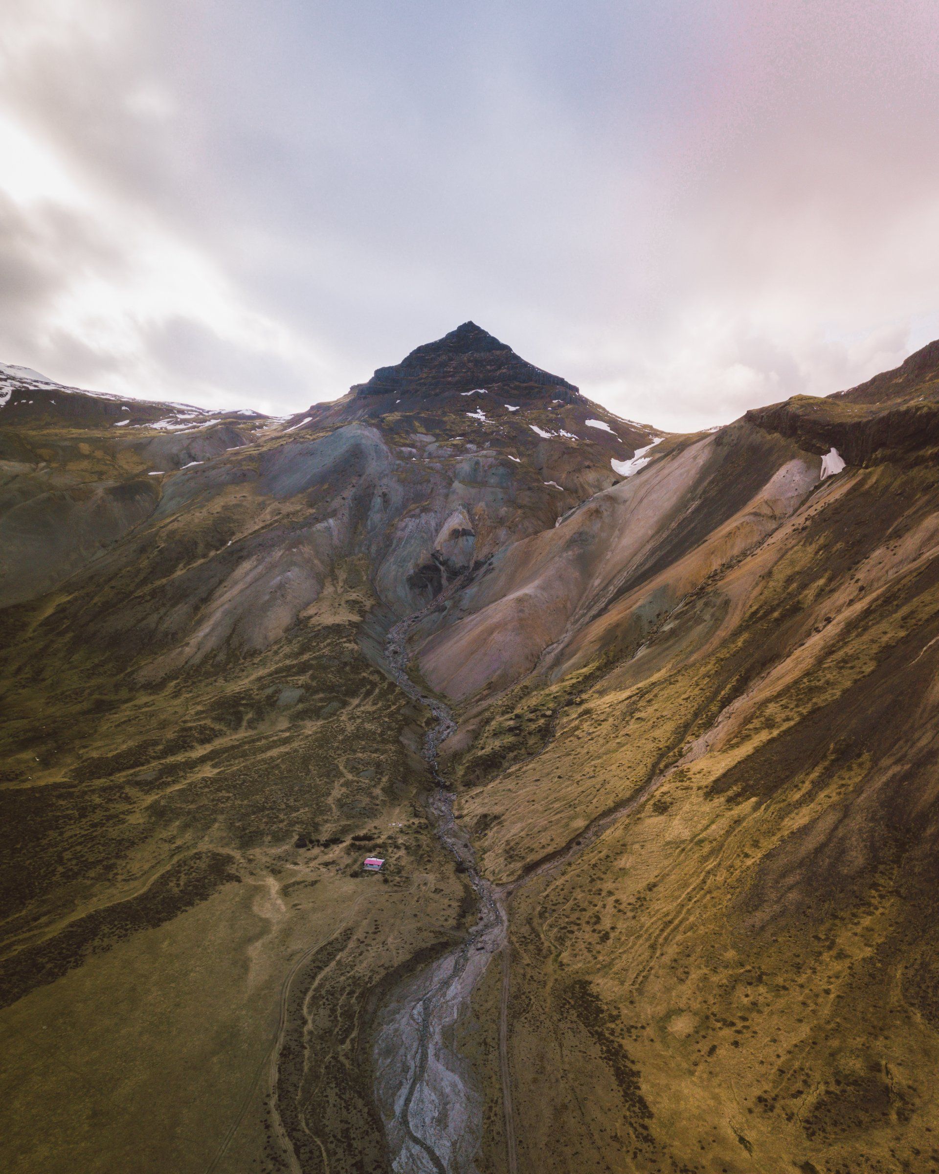 Berggipfel mit freiliegendem Fels, grasbewachsenen Hängen und einem Bach unter einem bewölkten Himmel.