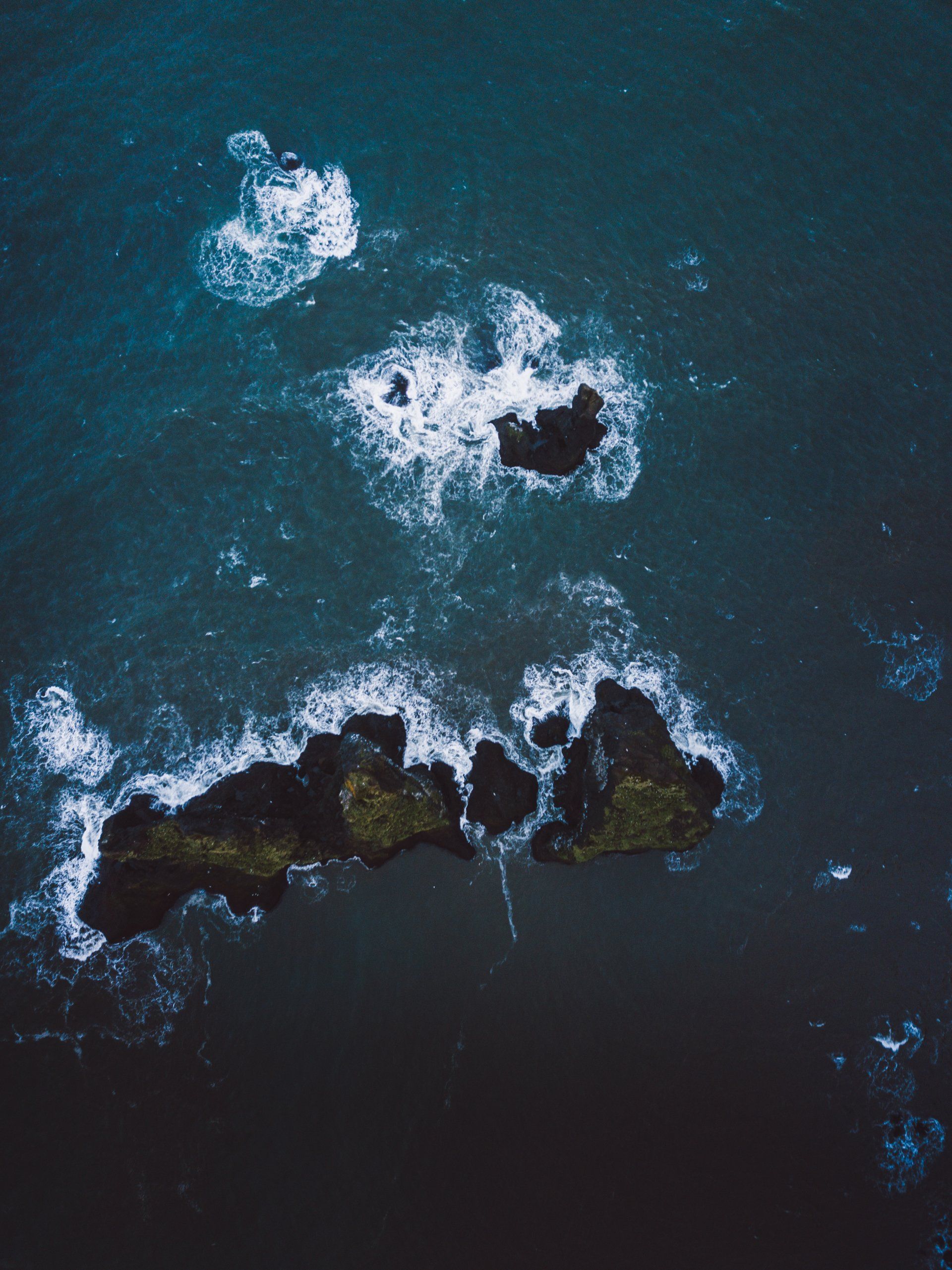 Luftaufnahme von felsigen Inseln im dunkelblauen Ozean, weiße, schäumende Wellen.