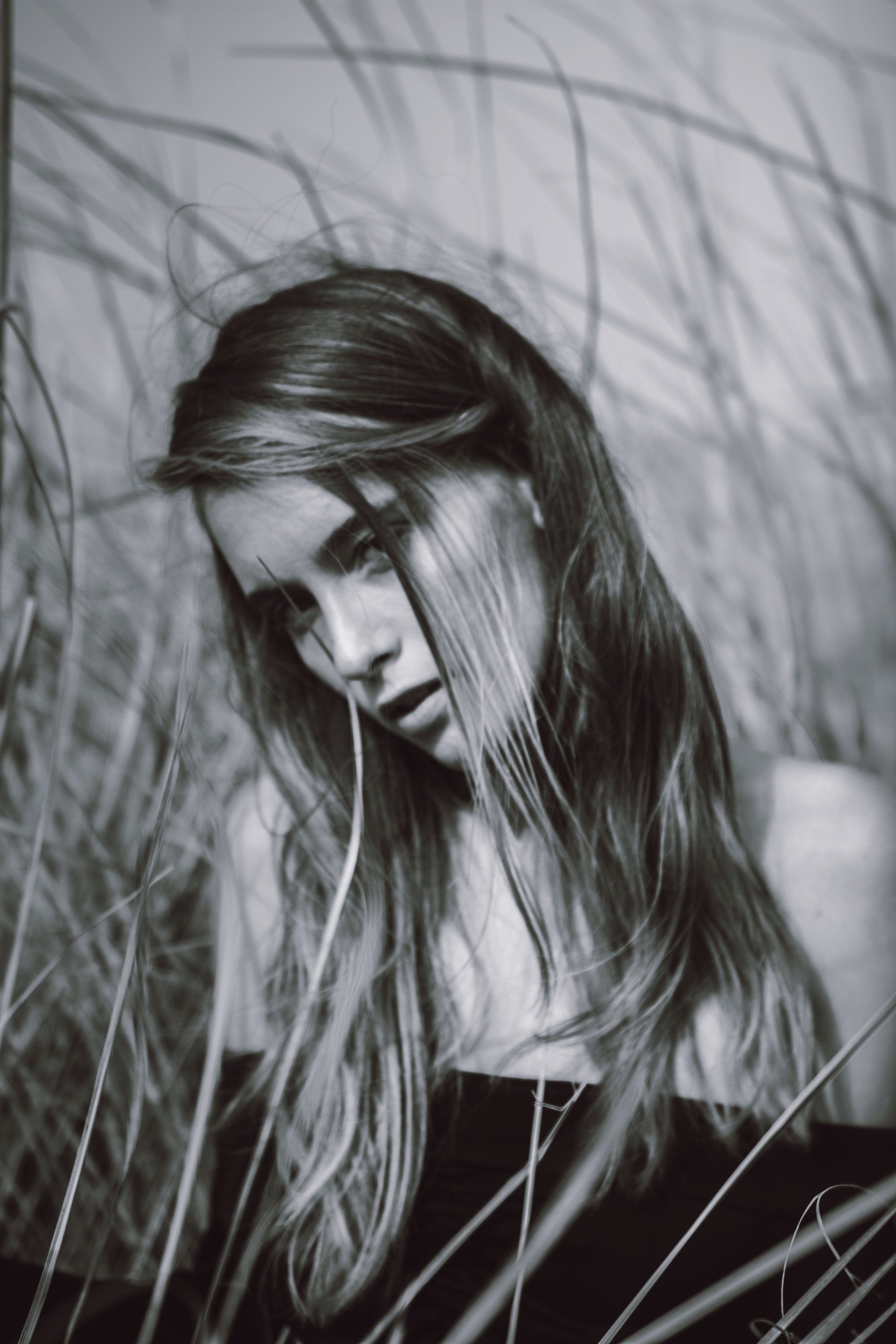 Frau mit langen Haaren blickt nach unten, teilweise verdeckt durch hohes Gras, in Monochrom.