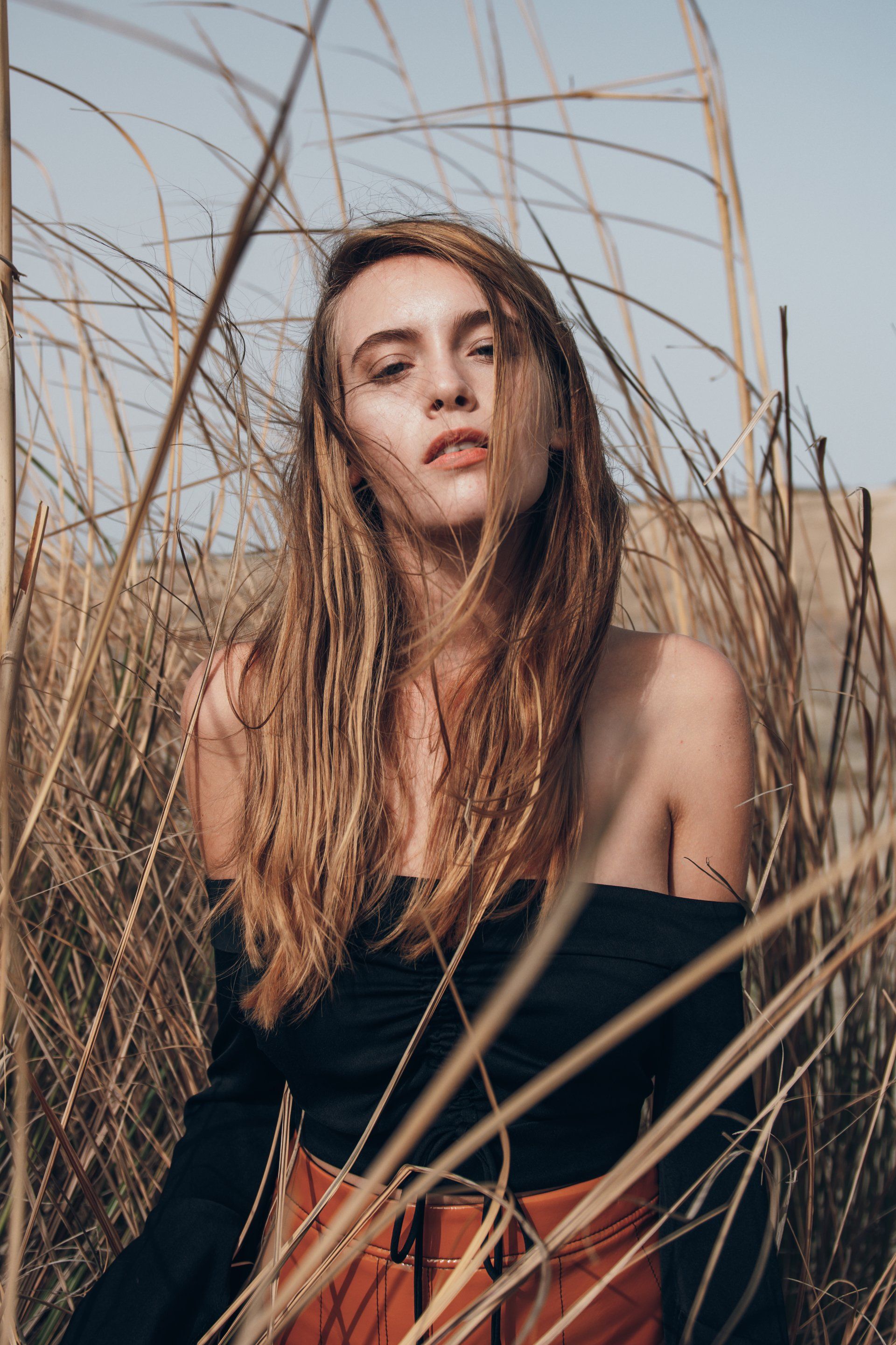 Frau mit langen Haaren, schwarzem Oberteil und roten Hosen, steht im hohen Gras und schaut in die Kamera.