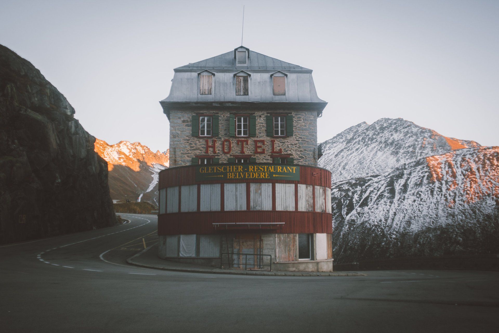 Hotelgebäude auf einem Gebirgspass, rotes Schild, graue Steinfassade, im Hintergrund schneebedeckte Gipfel.