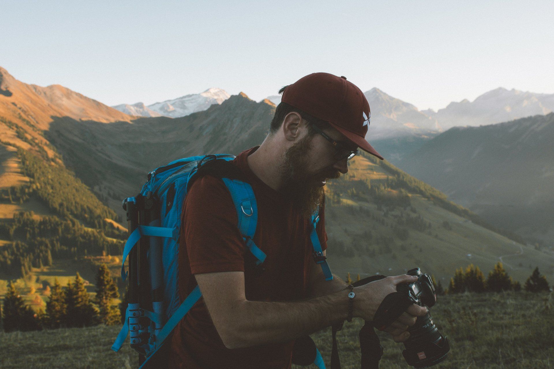 Mann mit Bart und roter Mütze, Rucksack, blickt in die Kamera vor Bergkulisse.