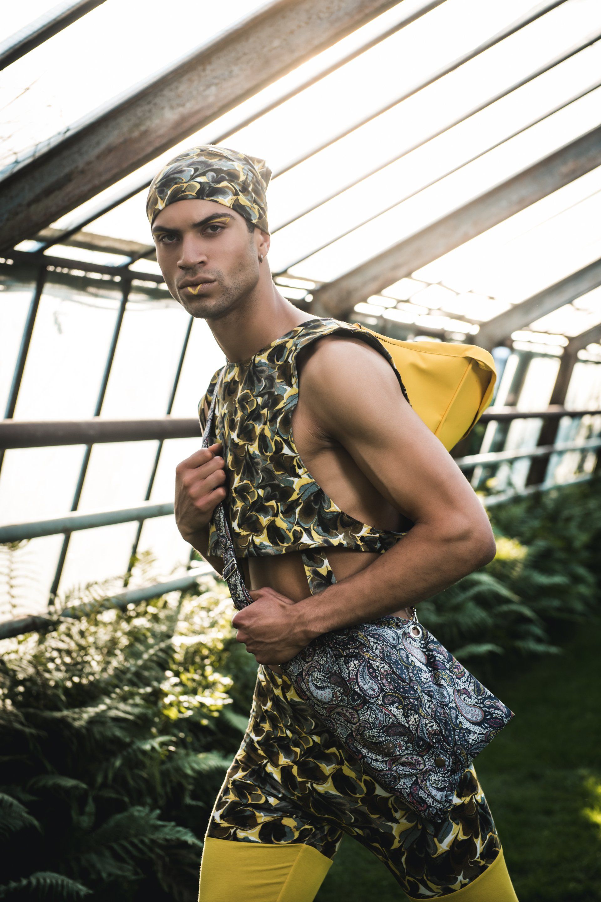 Mann in gemustertem Outfit posiert in einem Gewächshaus. Gelbe Leggings, Tasche.