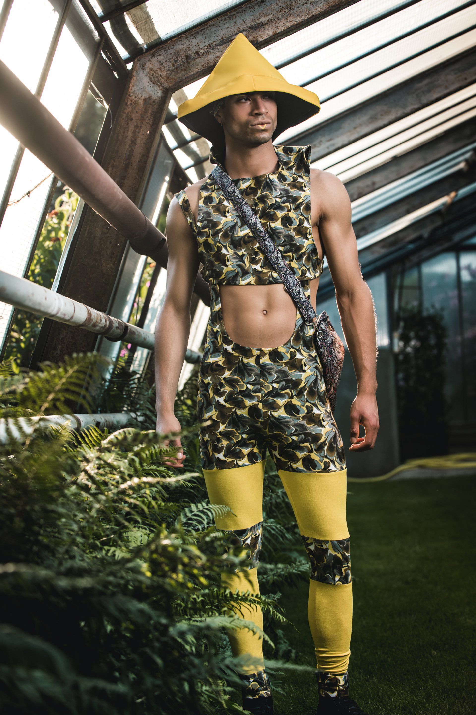 Mann in Tarnkleidung mit gelben Akzenten und Hut steht in einem Gewächshaus.