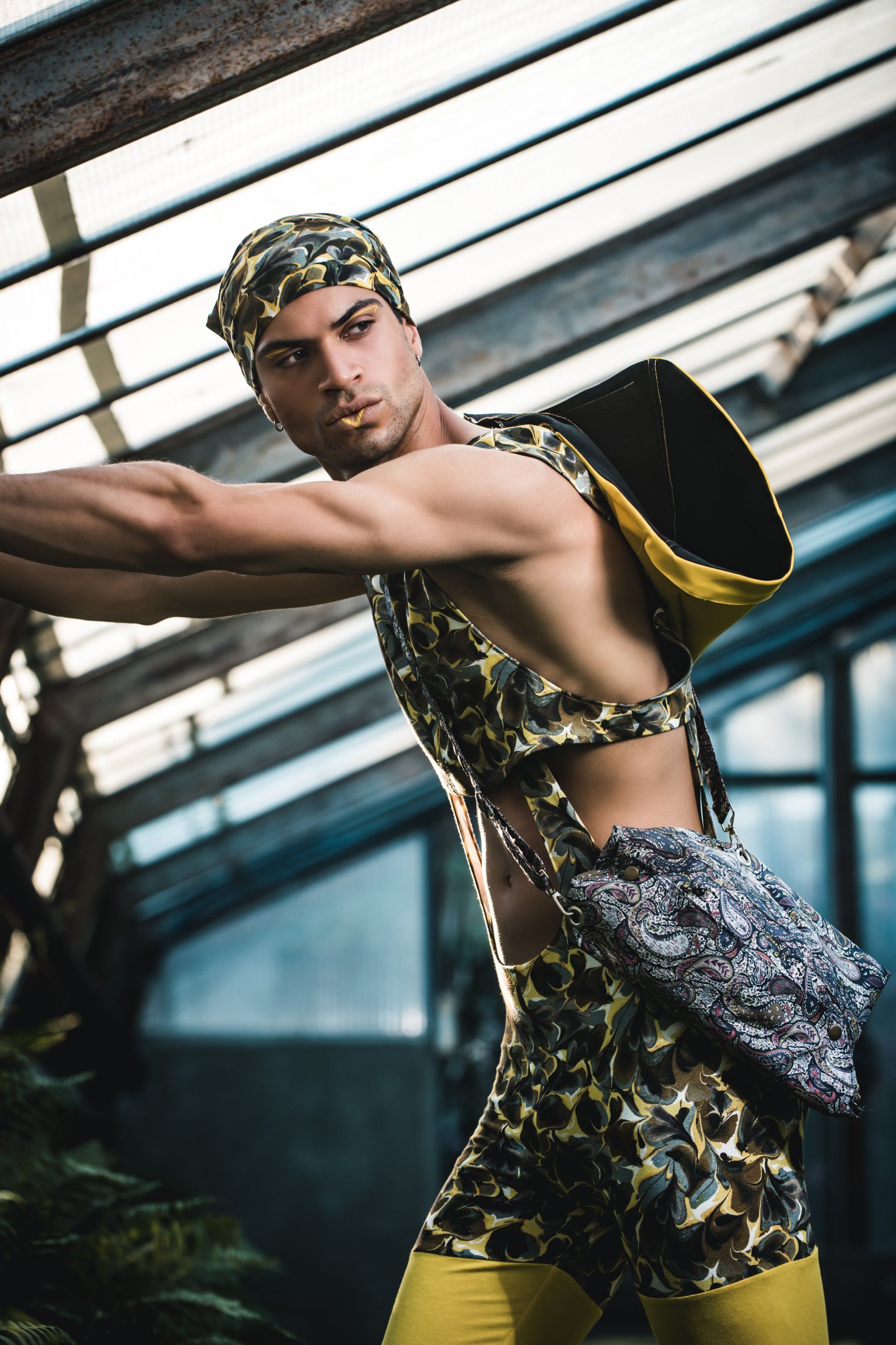 Mann in gemusterter Kleidung, lehnt mit Rucksack und Kopftuch in einem Gewächshaus.