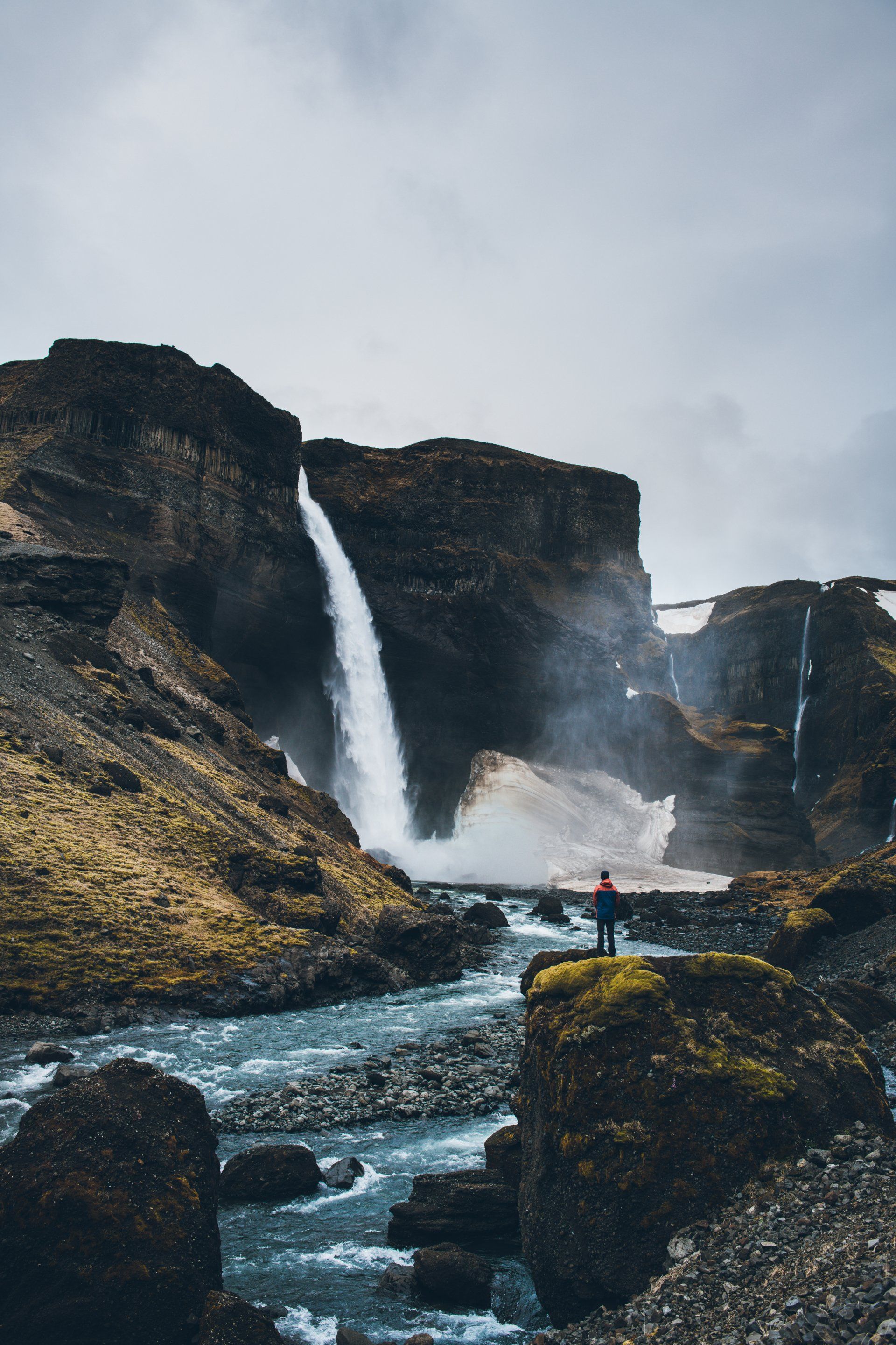 Person steht vor einem großen Wasserfall, der eine schroffe Klippe hinunterstürzt. Bewölkter Tag, darunter ein Fluss.