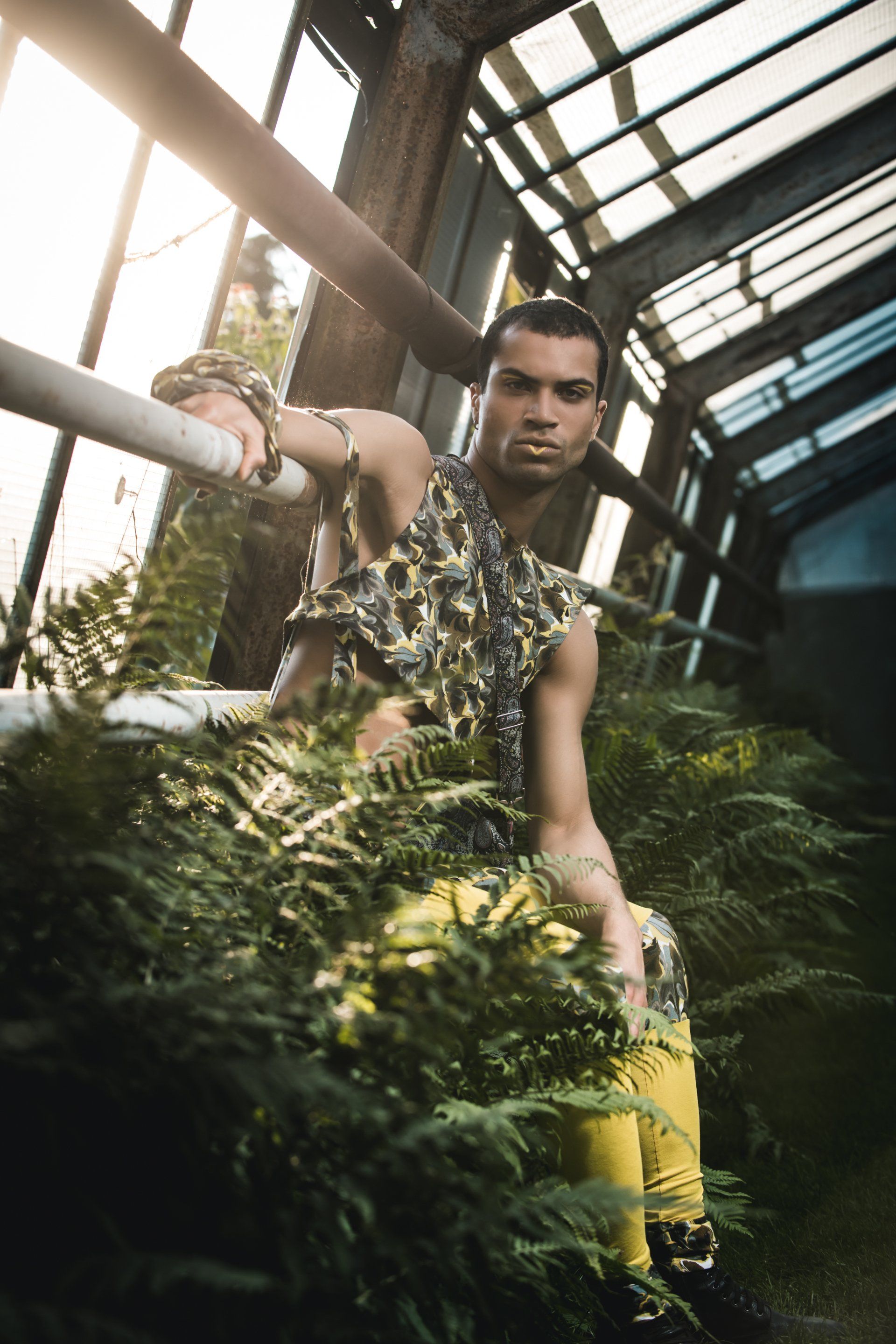 Mann in gemustertem Oberteil und gelber Hose posiert inmitten von Grün in einem Glasgewächshaus.
