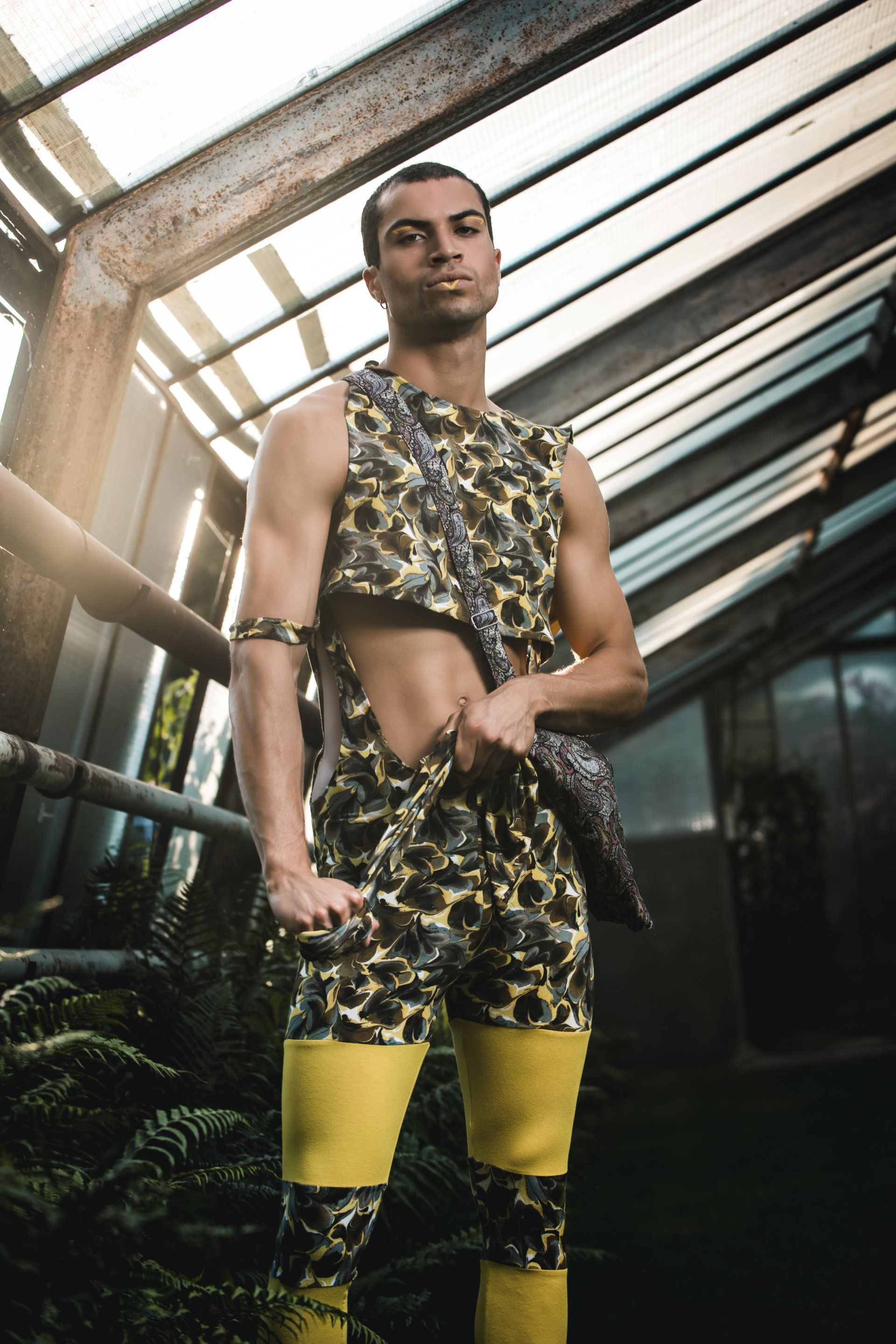 Mann im Tarnoutfit mit gelben Stiefeln und Armbinden posiert in einem Gewächshaus.