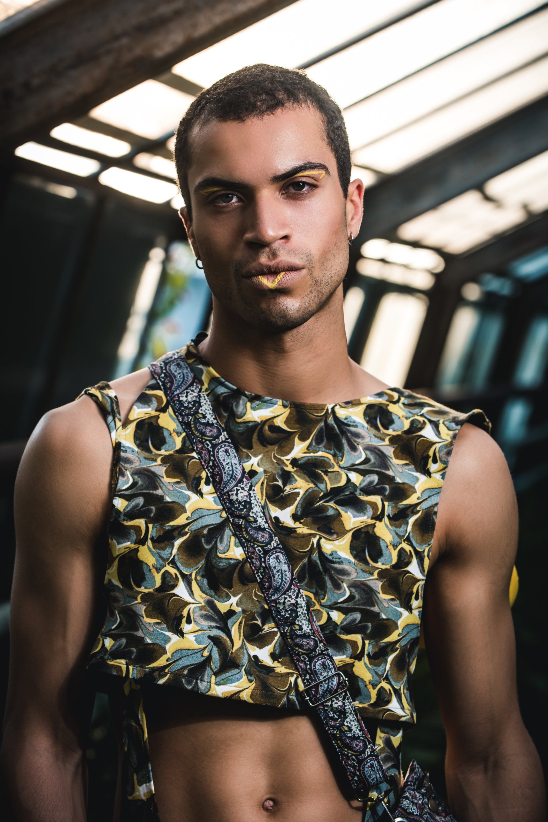 Eine farbige Person mit ernstem Blick, die ein gemustertes Oberteil und einen Träger trägt; Hintergrund mit Oberlicht.