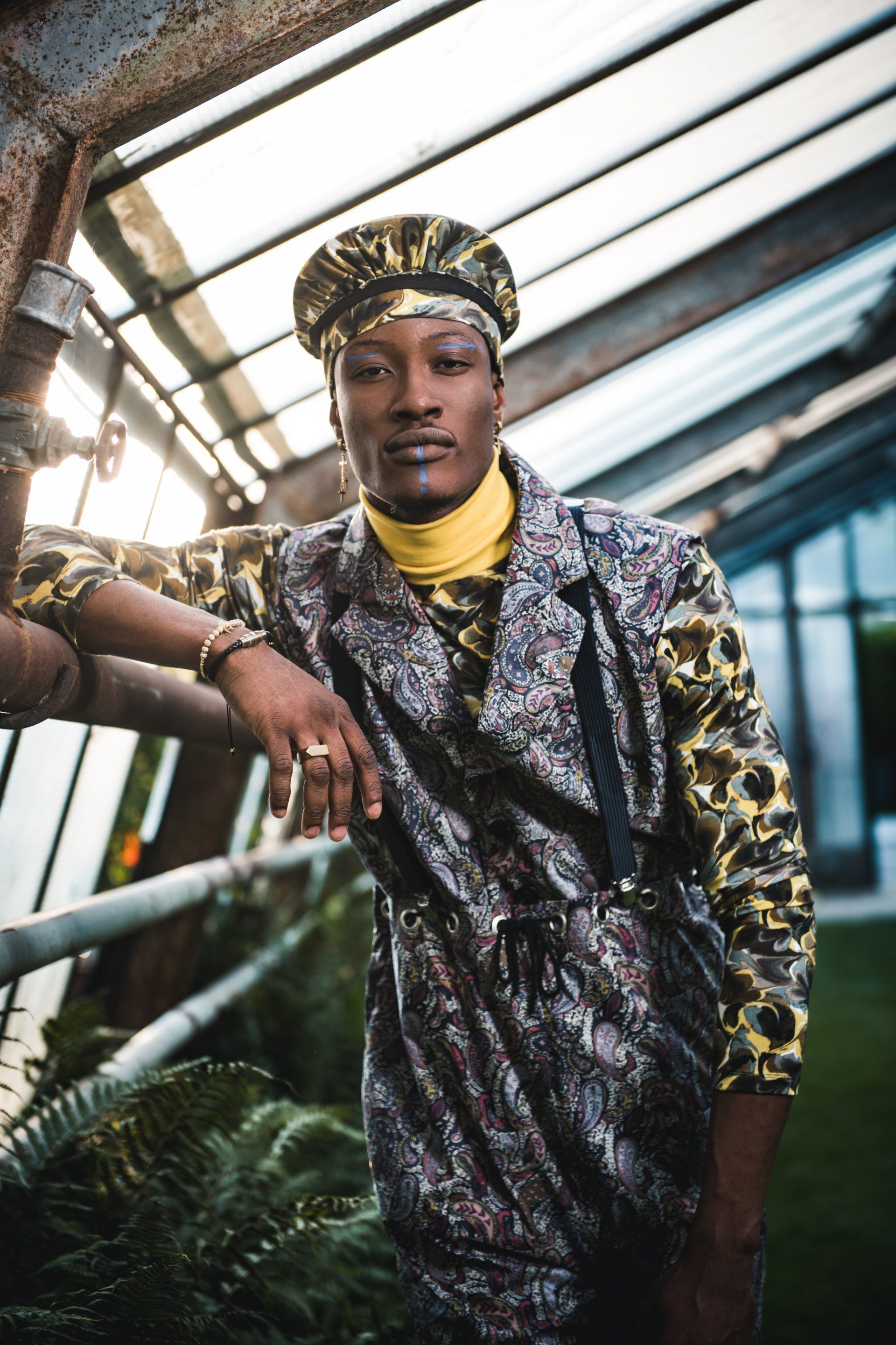 Mann in gemusterter Jacke und Hut lehnt an Metallkonstruktion im Gewächshaus.