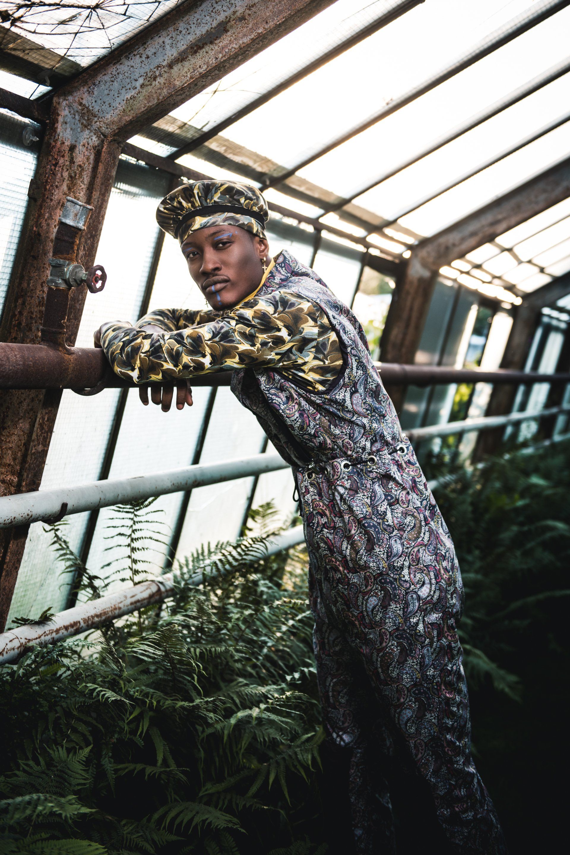 Frau in gemustertem Outfit lehnt in einem Gewächshaus an einem Metallrohr und blickt in die Kamera.