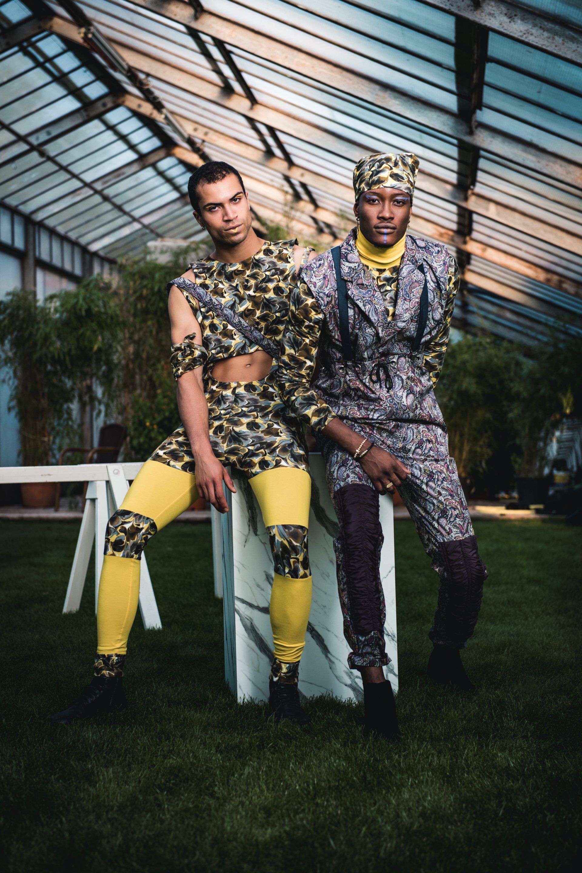 Zwei Personen in gemusterten Outfits posieren in einem Glasgewächshaus.