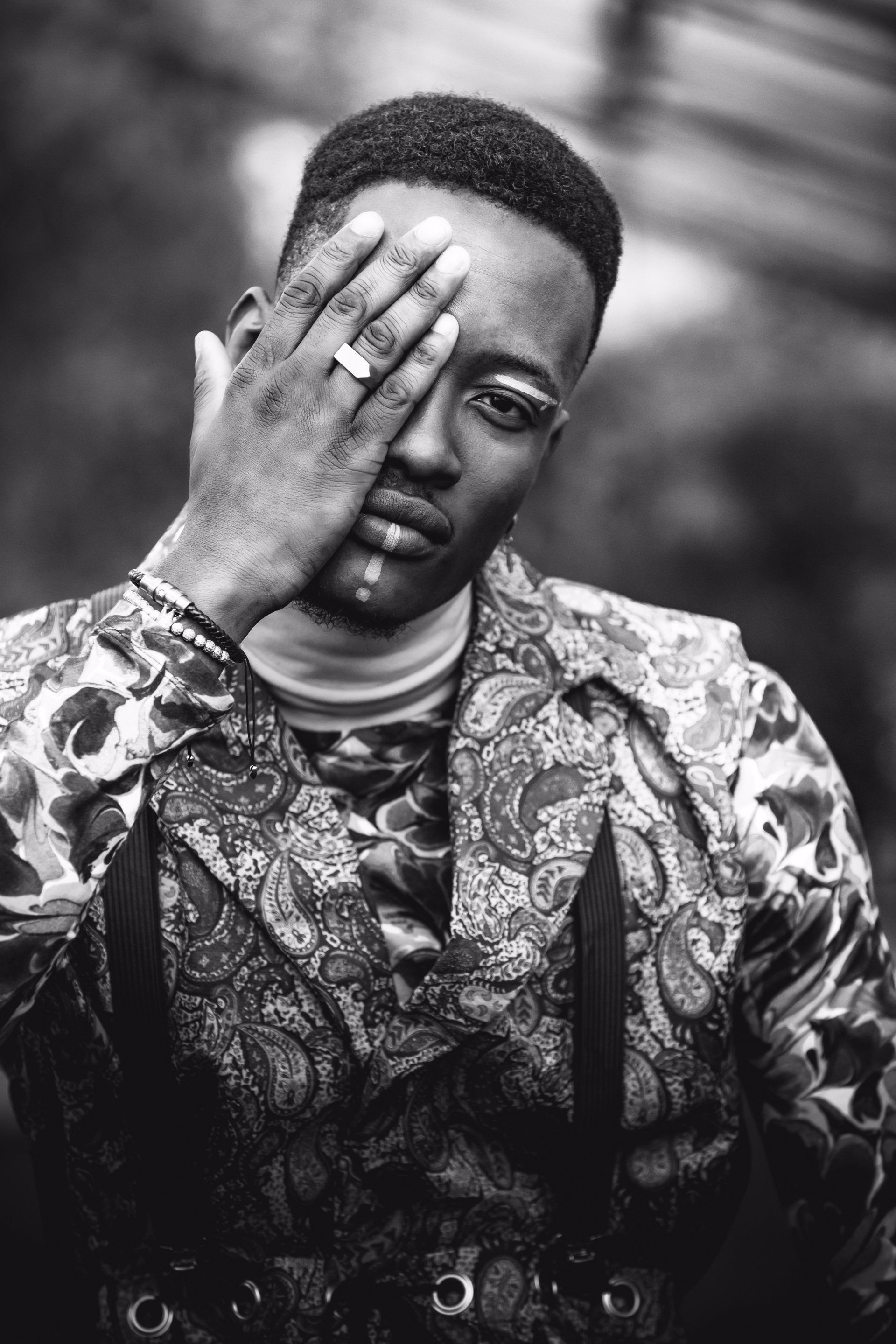 Schwarz-Weiß-Porträt eines Mannes, der ein Auge bedeckt. Er trägt eine gemusterte Jacke und einen Ring.