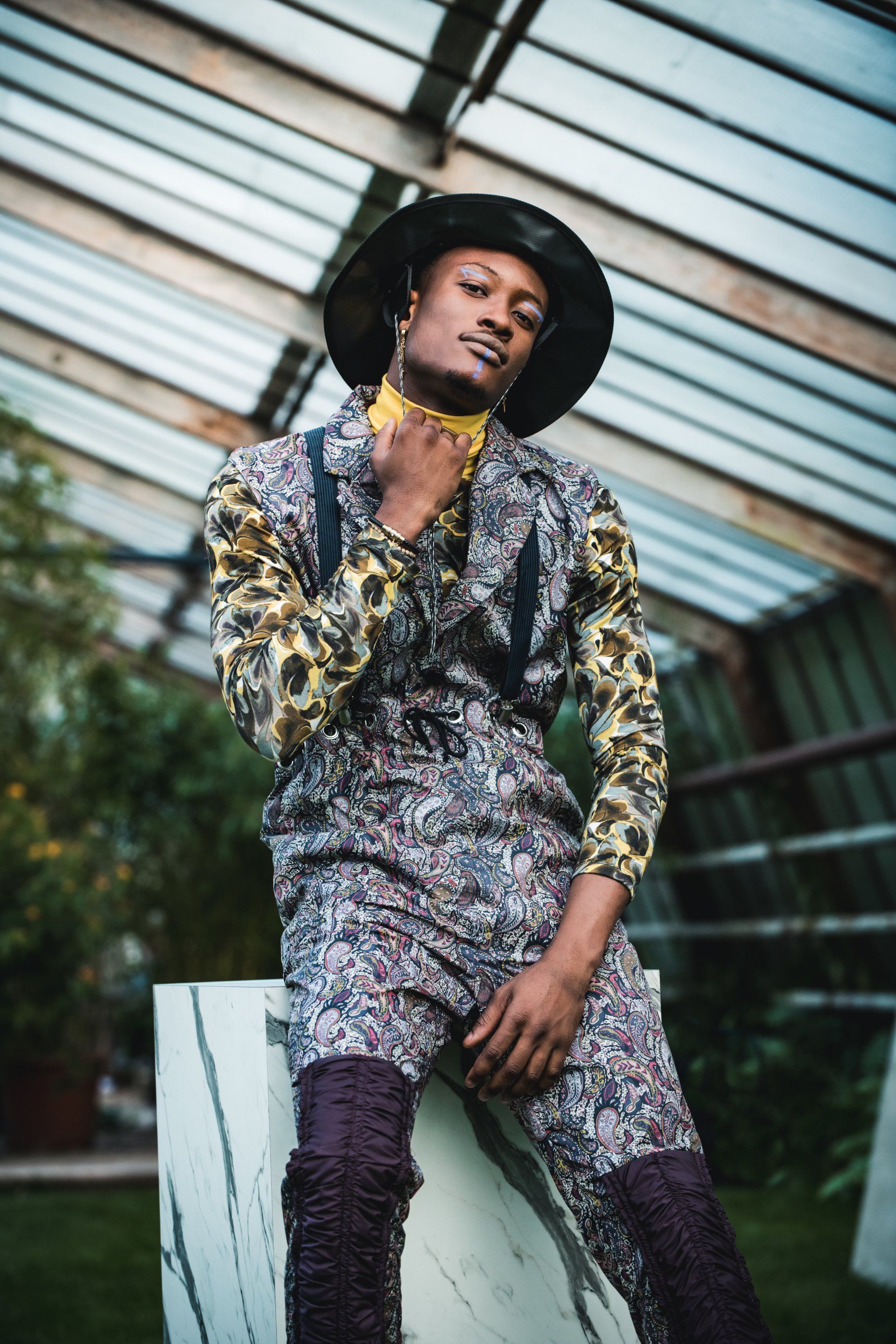 Eine Person in gemustertem Overall und Hut posiert in einer Struktur mit Glasdach und blickt in die Kamera.