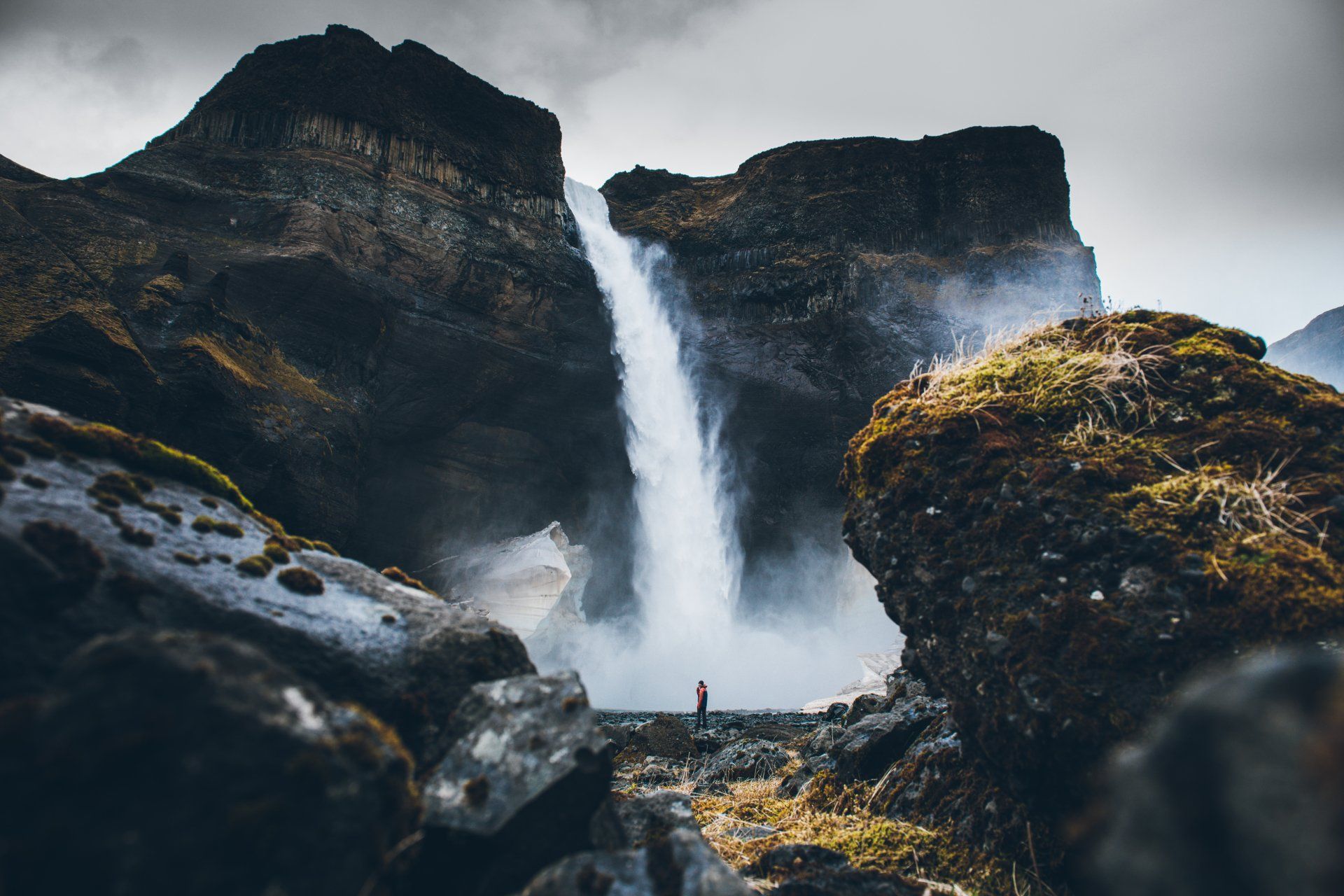 Wasserfall stürzt zwischen Felsklippen hinab, eine winzige Gestalt steht am Fuße. Stimmungsvoller, bewölkter Himmel.