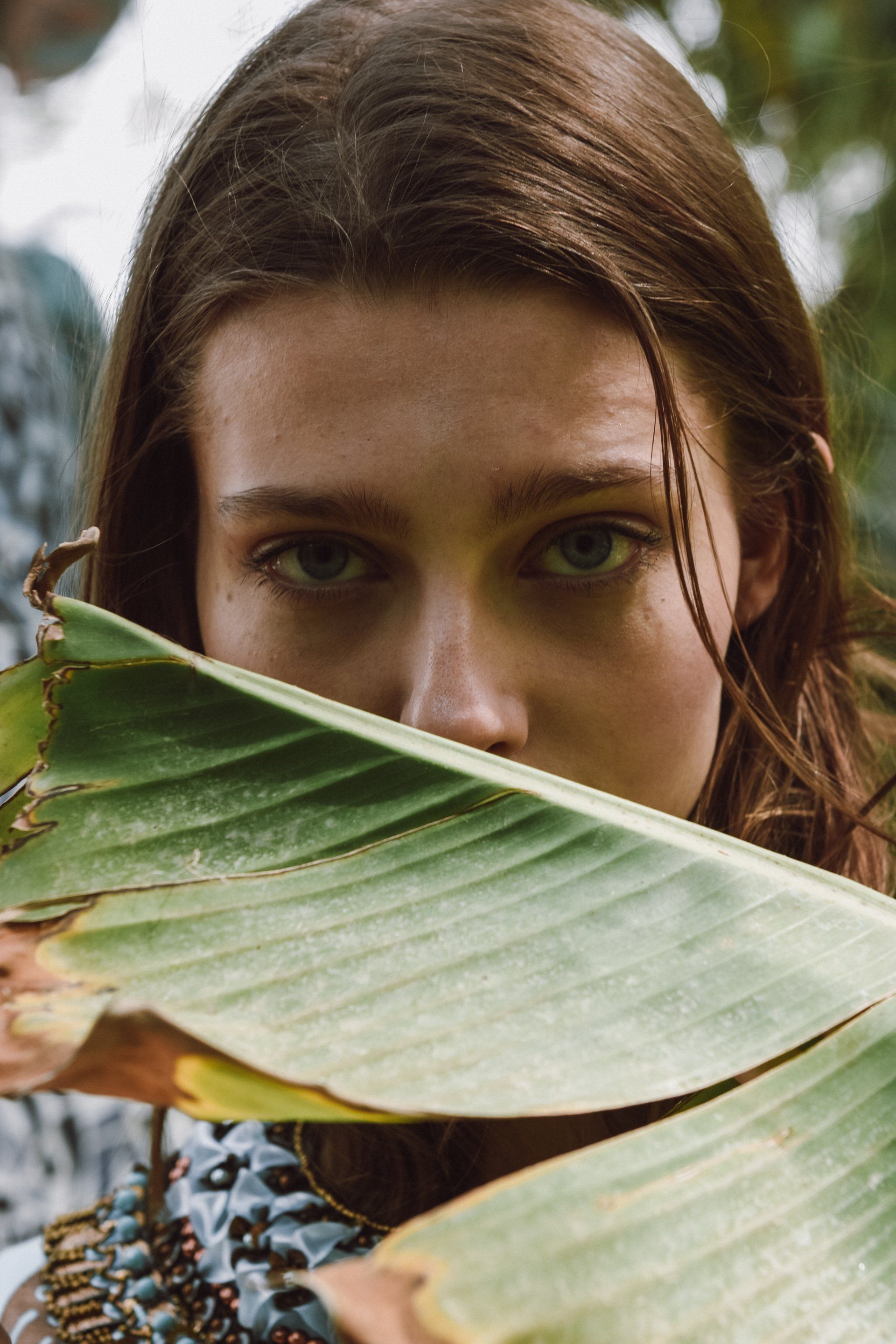 Frau blickt durch ein großes grünes Blatt; sanftes Licht, braunes Haar, konzentrierter Ausdruck, Umgebung im Freien.