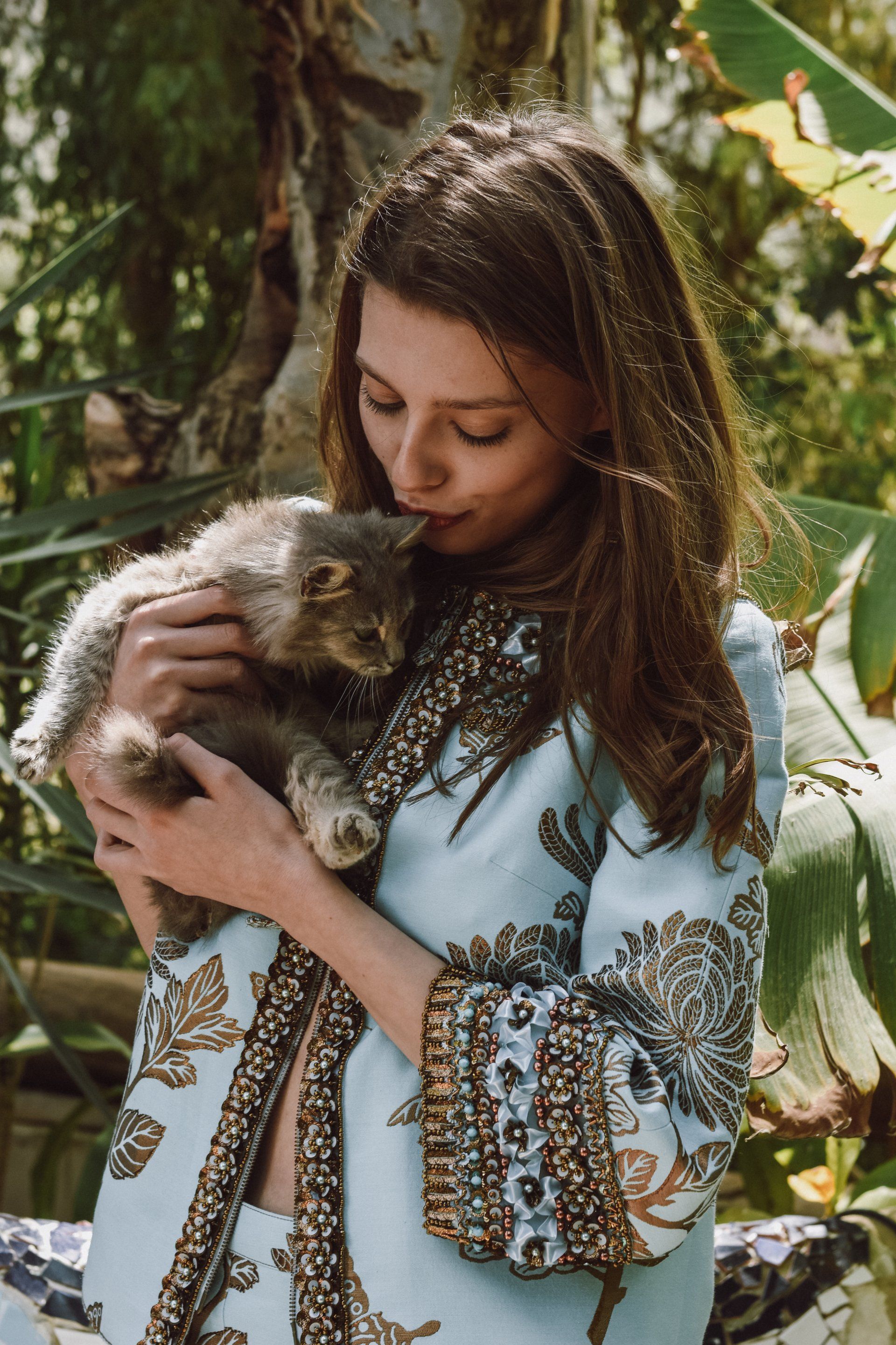 Frau in blau verzierter Jacke hält und küsst im Freien eine flauschige graue Katze.