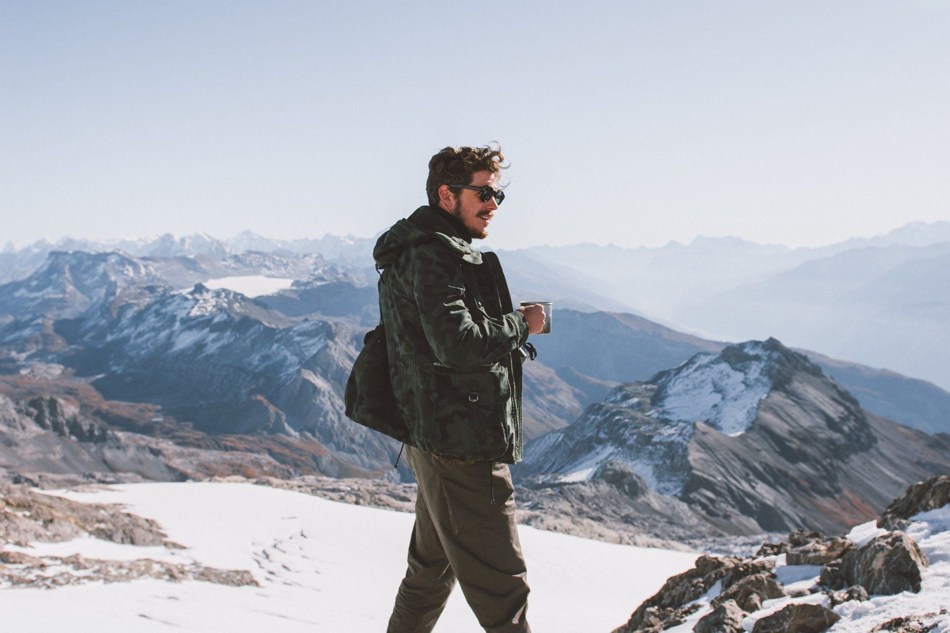 Mann mit Sonnenbrille und Jacke, blickt auf schneebedeckte Berge und hält einen Becher.