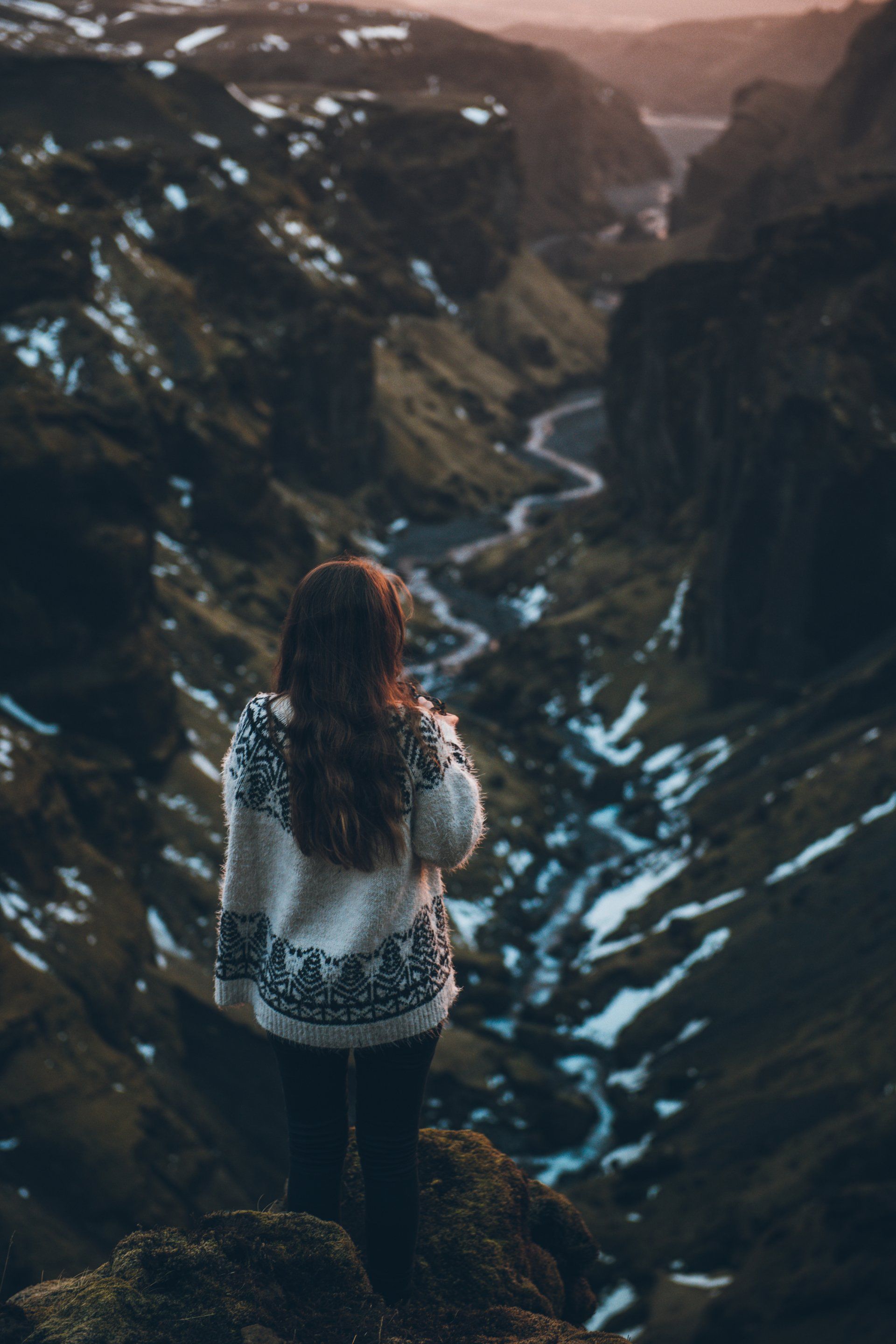 Frau im gemusterten Pullover überblickt ein gewundenes Flusstal, Schneeflecken auf grünen Hängen, dramatische Landschaft.