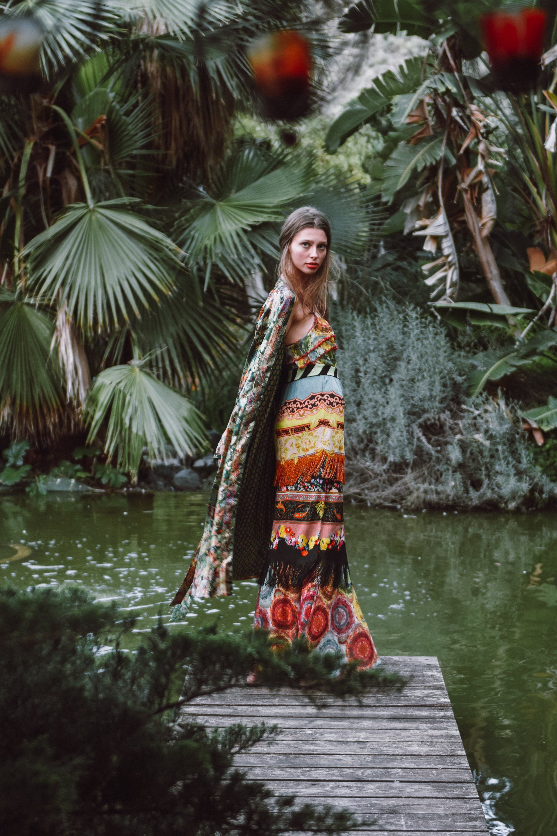Frau auf einem Holzsteg, in einem bunten Kleid und Umhang, in einem üppigen, grünen Garten am Wasser.